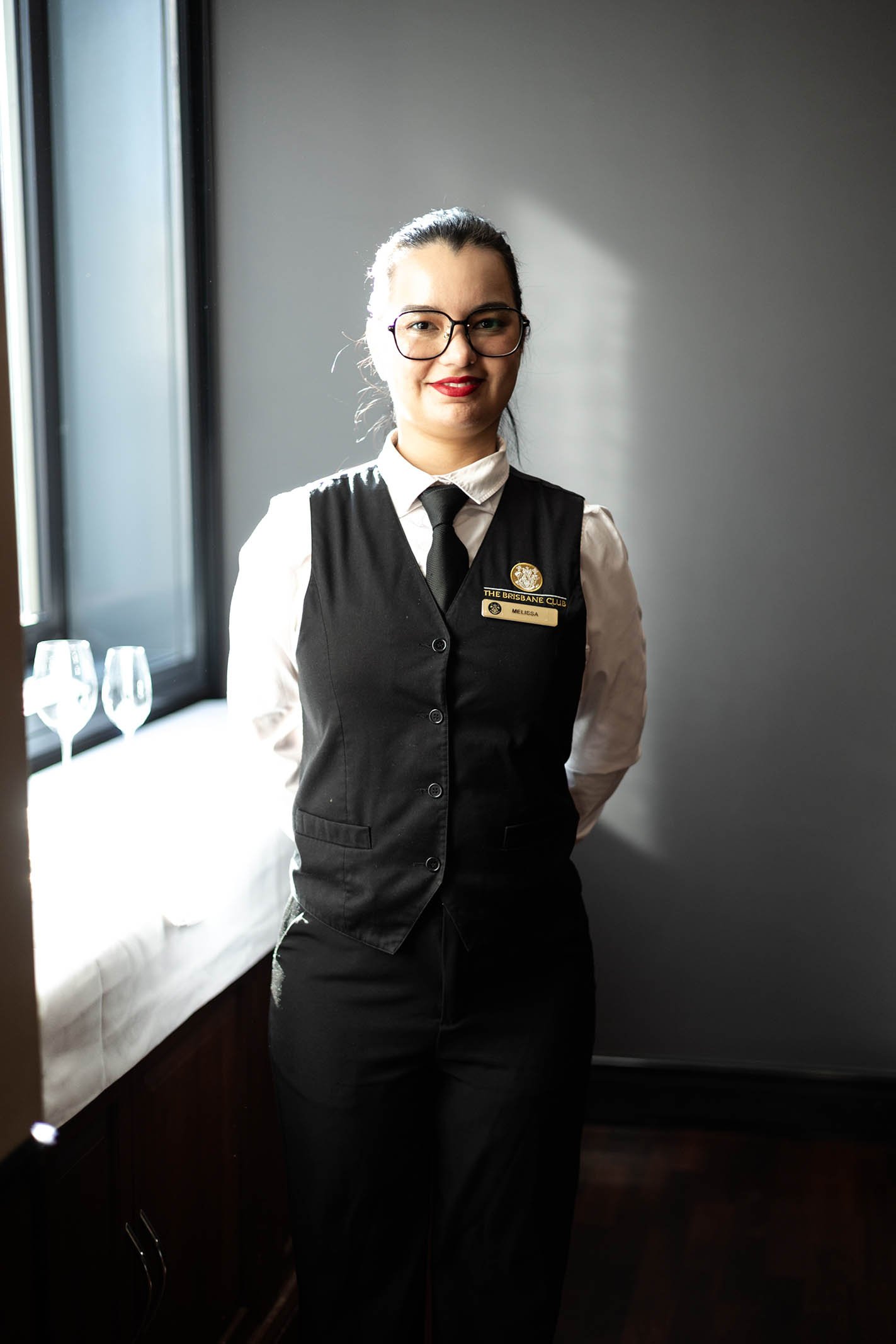 A woman with dark hair, glasses, and red lipstick standing indoors near a window, wearing a black vest with a name tag and a white shirt underneath, with wine glasses on the windowsill behind her.