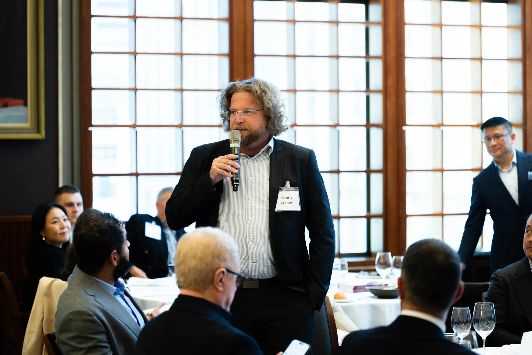 A man with curly hair, glasses, and a beard is holding a microphone and speaking at a formal event with attendees seated at tables. He is wearing a black blazer and a name tag that reads "Grant Mason". The setting is a well-lit room with large window