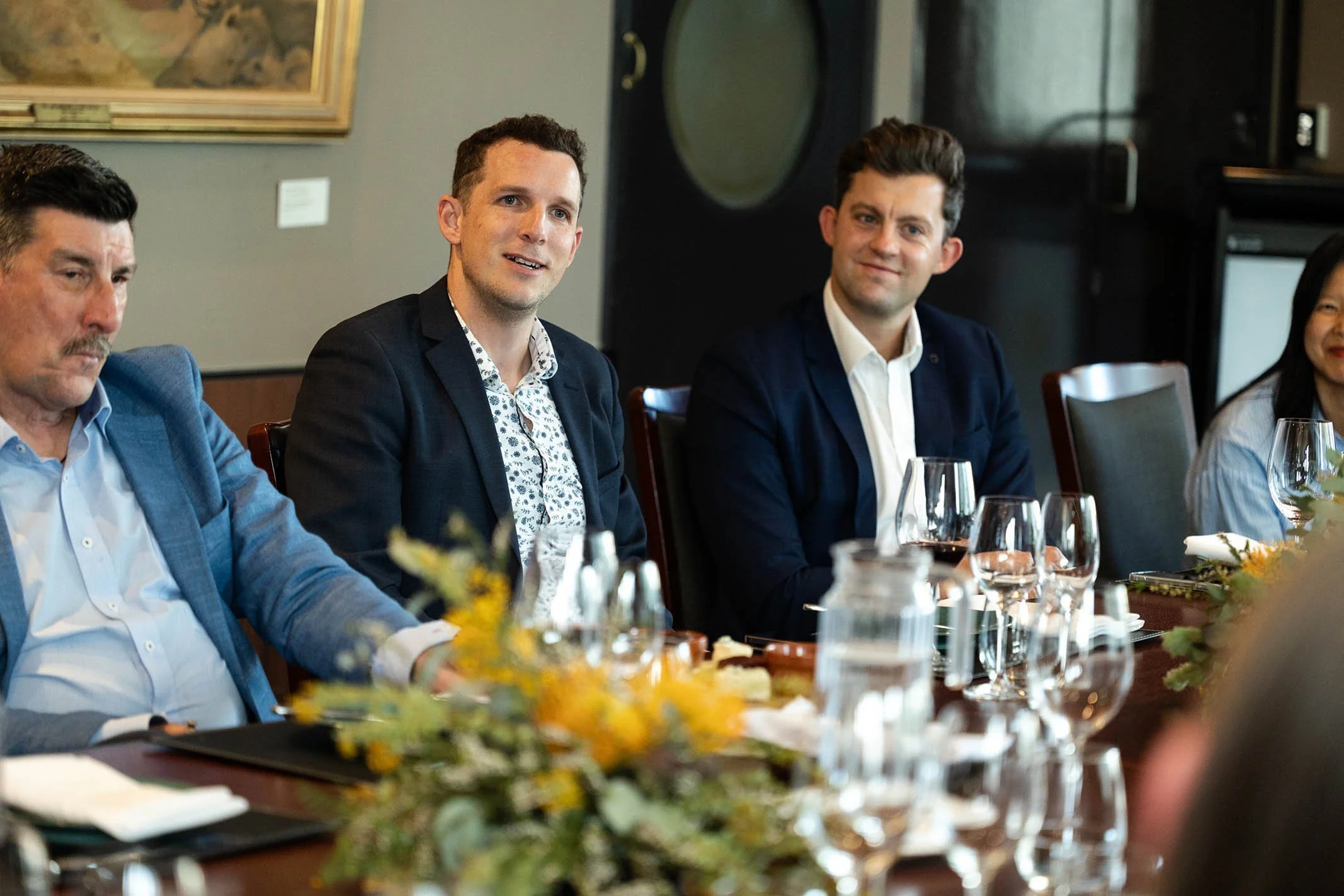 A group of five people sitting at a dining table during a formal event. The table is decorated with a floral centerpiece, and there are wine glasses and water pitchers. The individuals are dressed in business attire, engaged in conversation, with a f