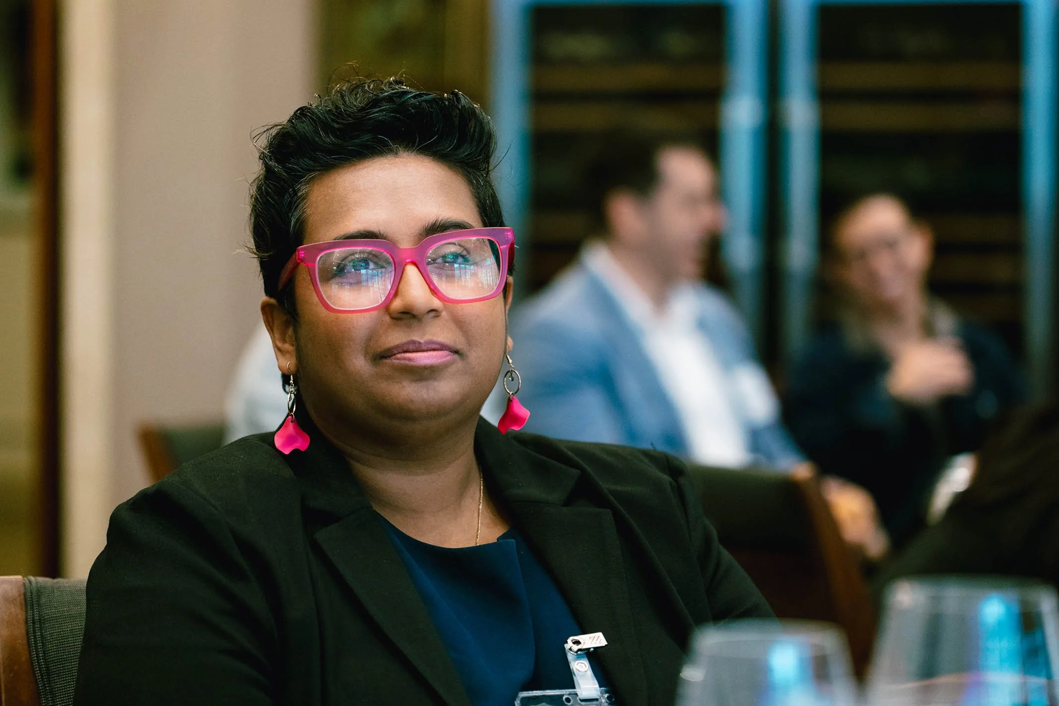 A woman with short black hair, wearing pink glasses and pink earrings, sits at a table during an event.