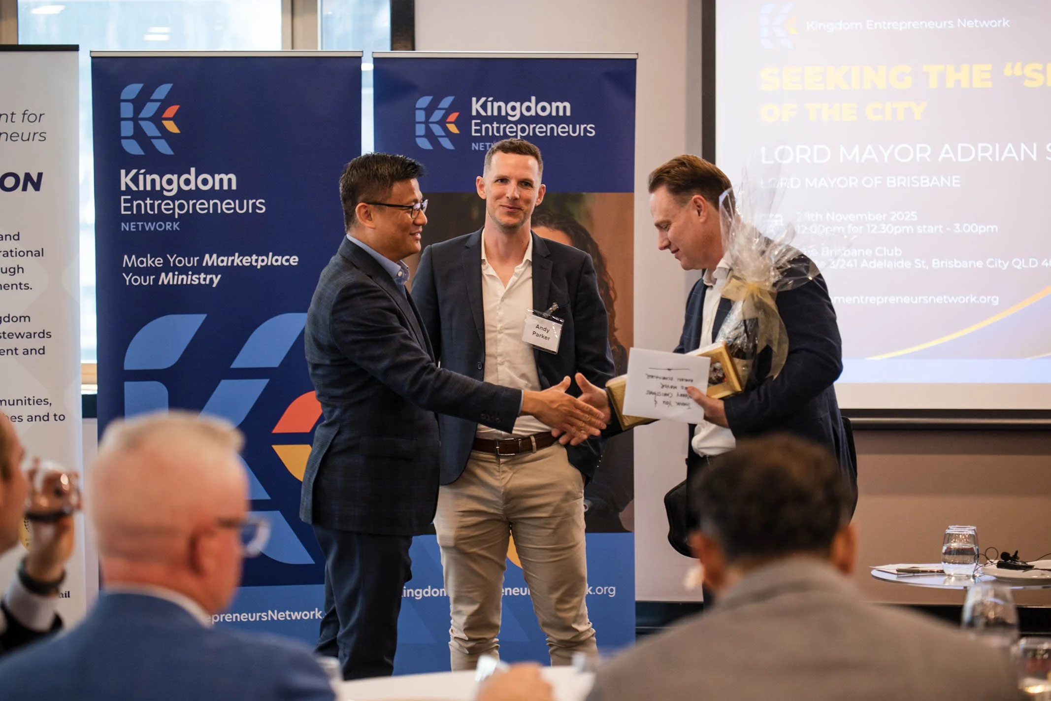 A man in a suit shaking hands with another man holding a gift, with a third man observing, at a professional event organized by Kingdom Entrepreneurs Network.