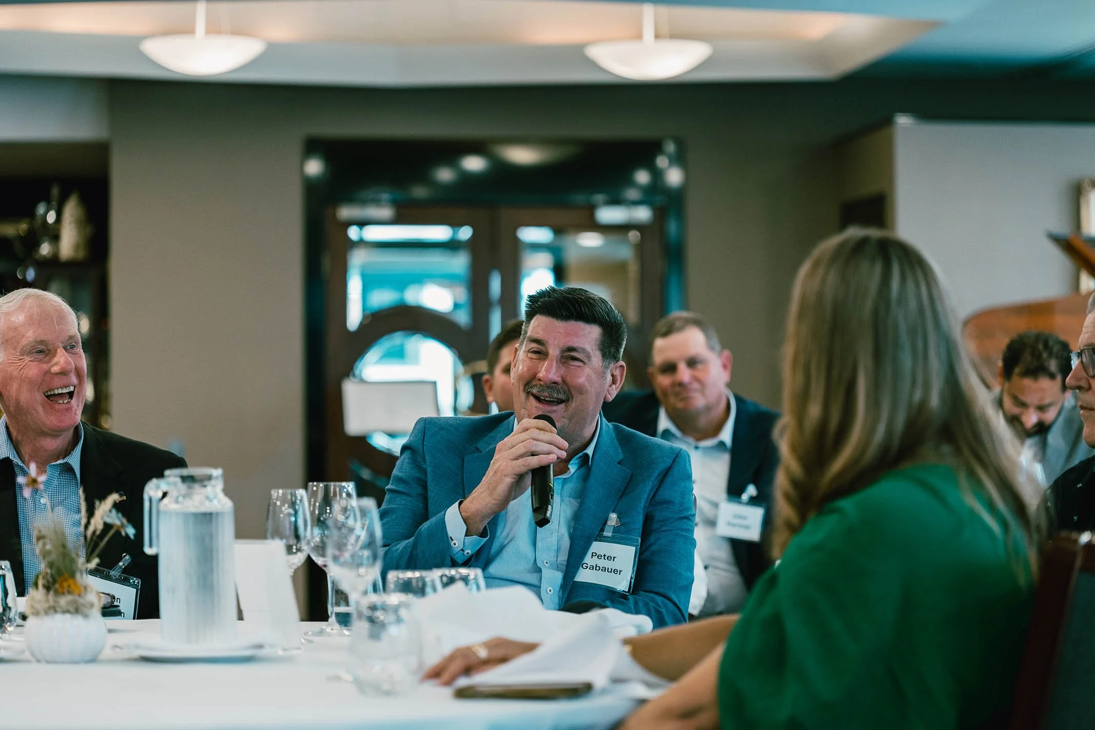 A man in a grey suit is speaking into a microphone at a business conference, surrounded by other attendees at a round table.