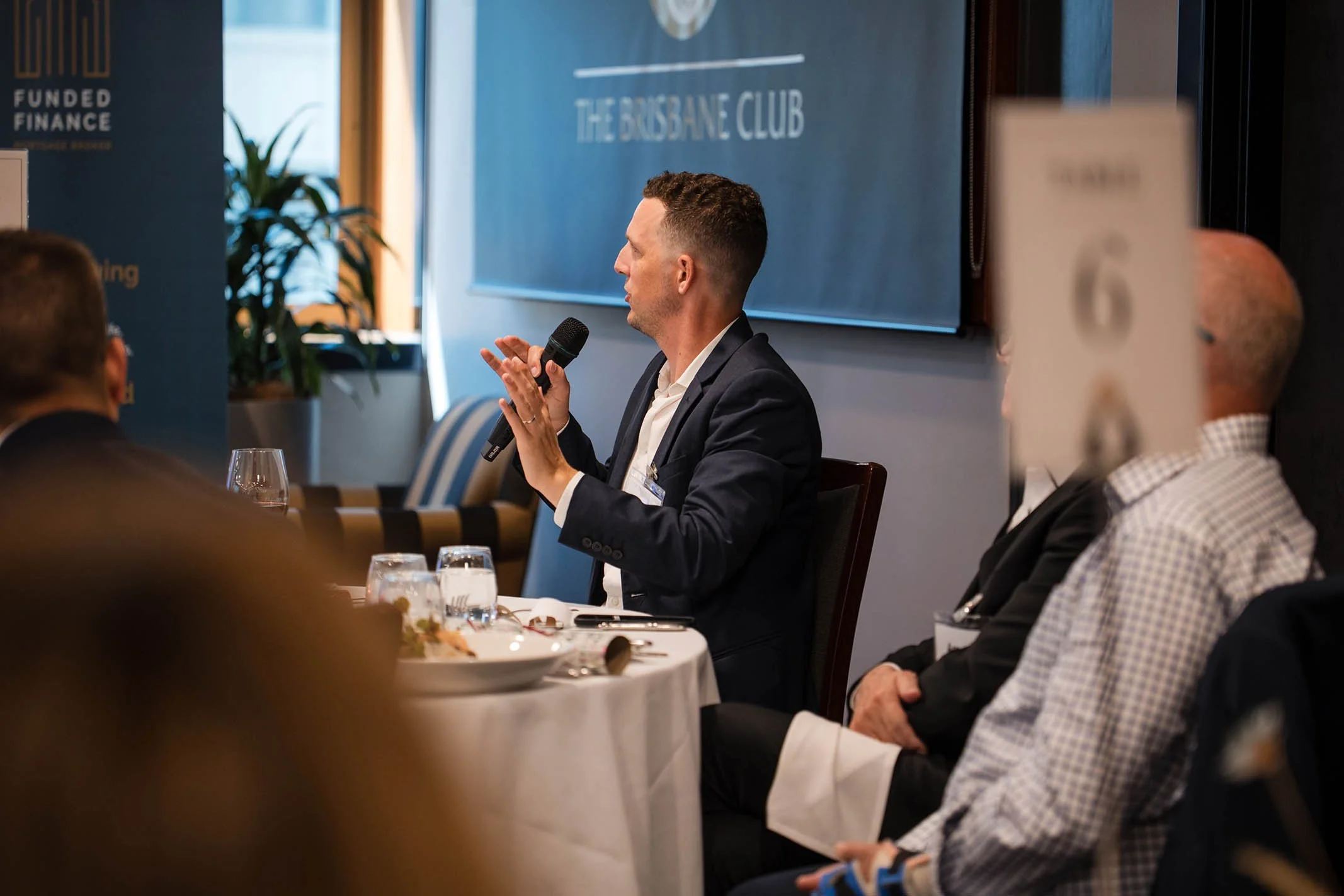 A man in a dark suit speaking into a microphone at a formal event, seated at a round table with glasses of water and plates, with a large screen displaying "The Brisbane Club" in the background.