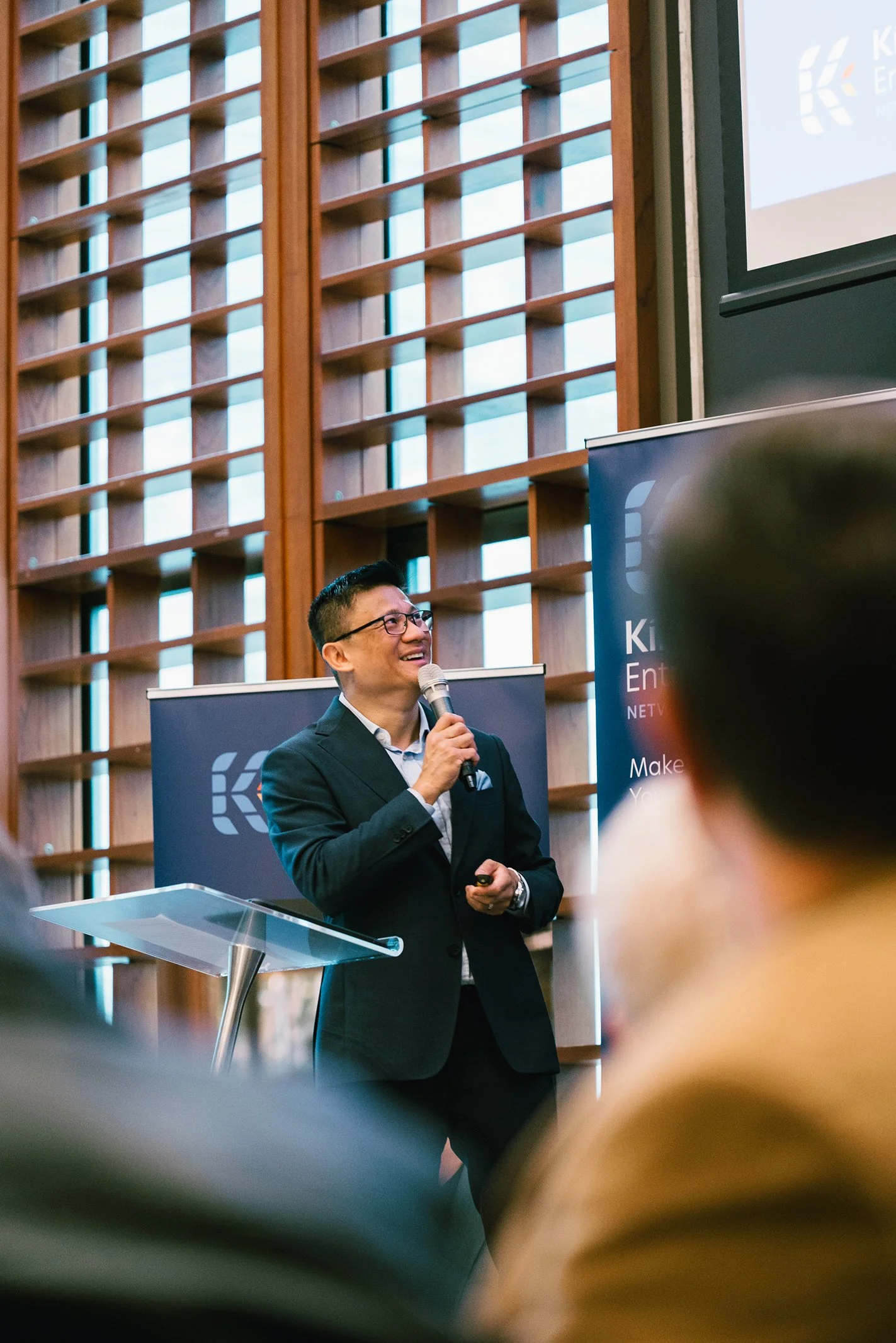 Man in a suit giving a presentation with a microphone in front of a wooden wall and a large screen at the event.