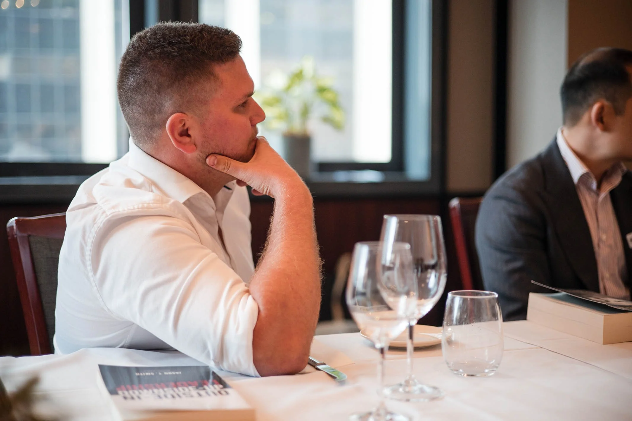 Man in a white shirt sitting at a table in a restaurant, with his hand on chin, near wine glasses and a book.