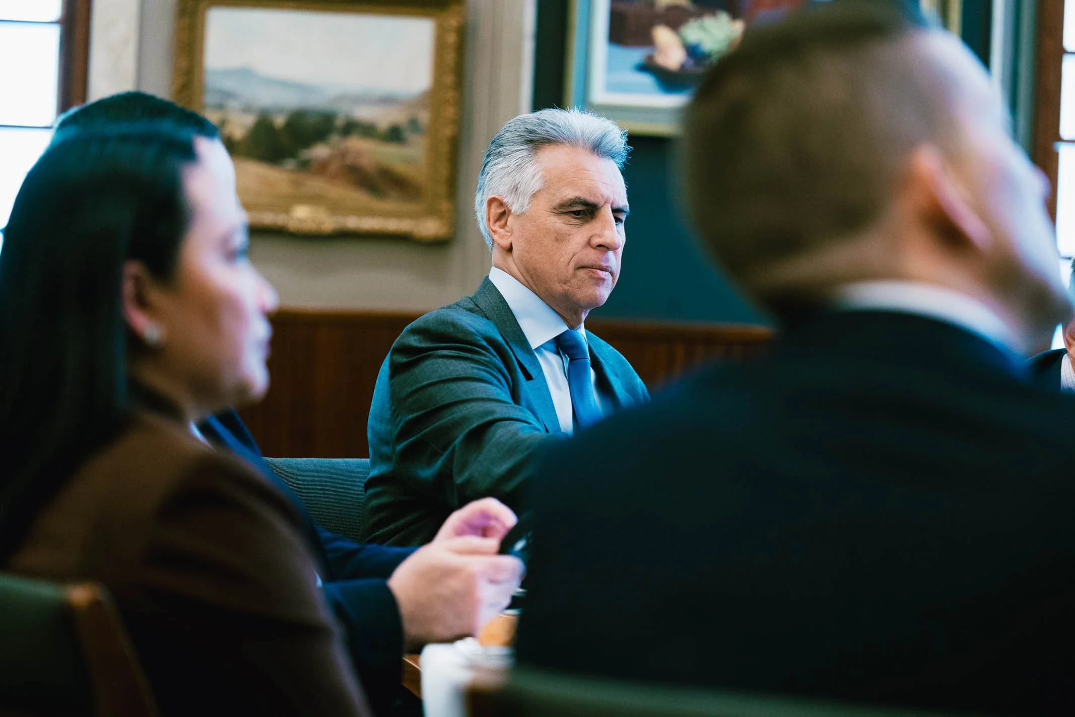 A man with gray hair and a serious expression, dressed in a suit and tie, sitting at a table with other people in a formal meeting or conference room with paintings on the wall.
