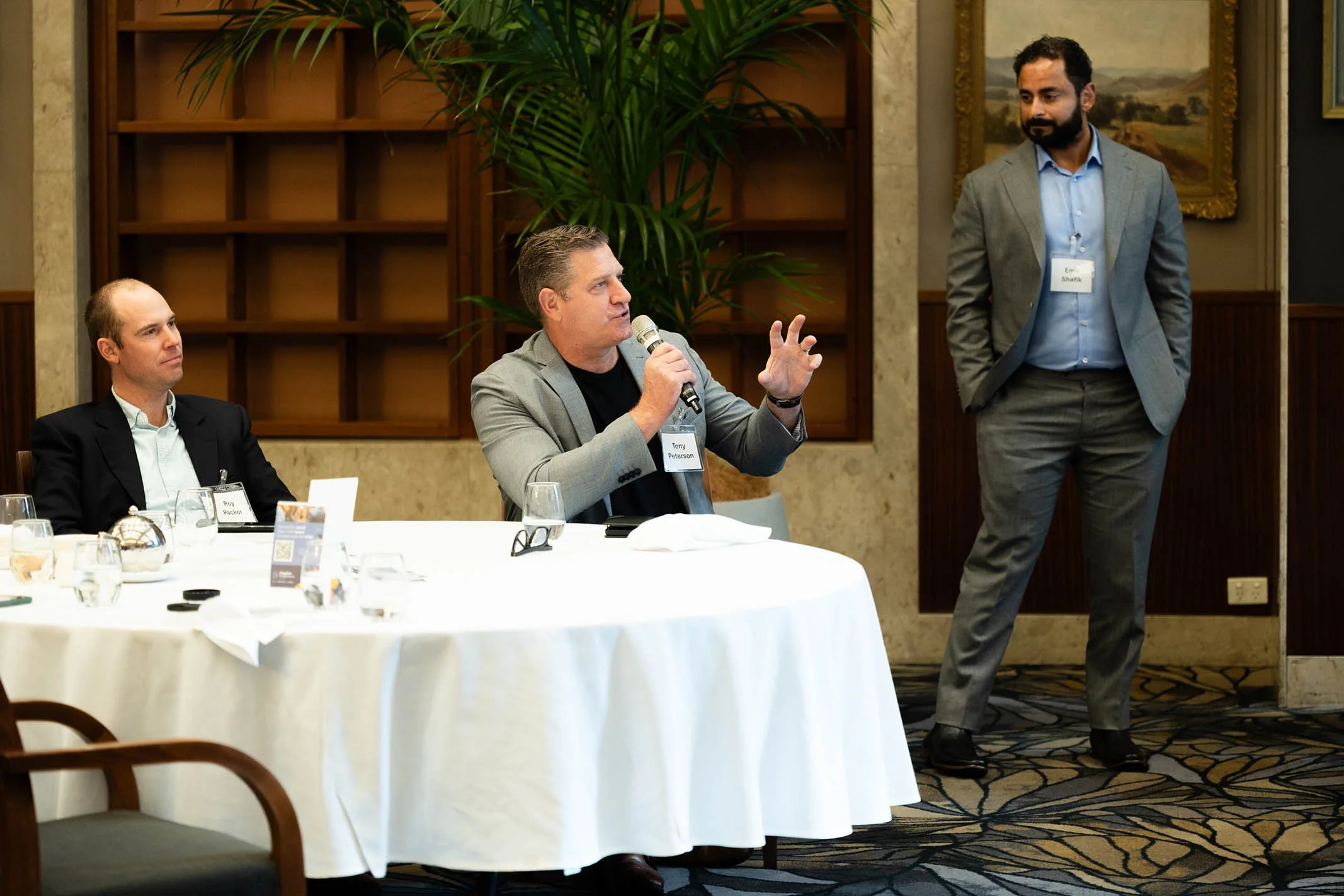 Three men in suits participating in a conference or meeting. One man is sitting at a round table, looking forward. Another man is seated, looking at the speaker. The third man is standing with his arms crossed, listening to the person with the microp