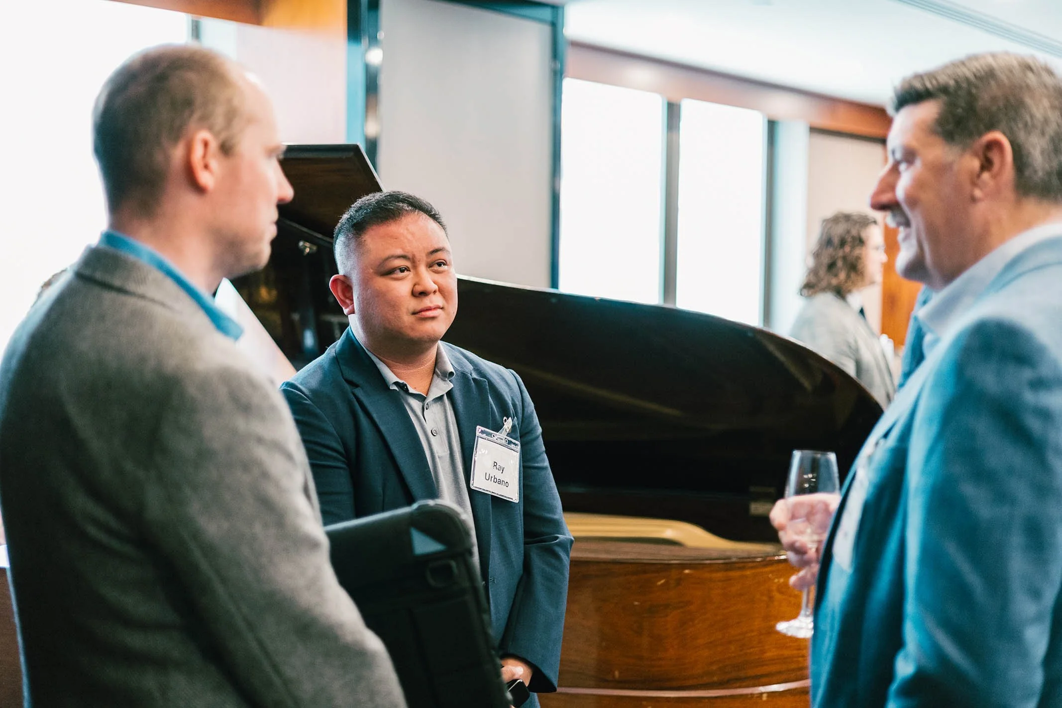 Three men in professional attire engaging in a conversation at a social event, with a grand piano in the background.