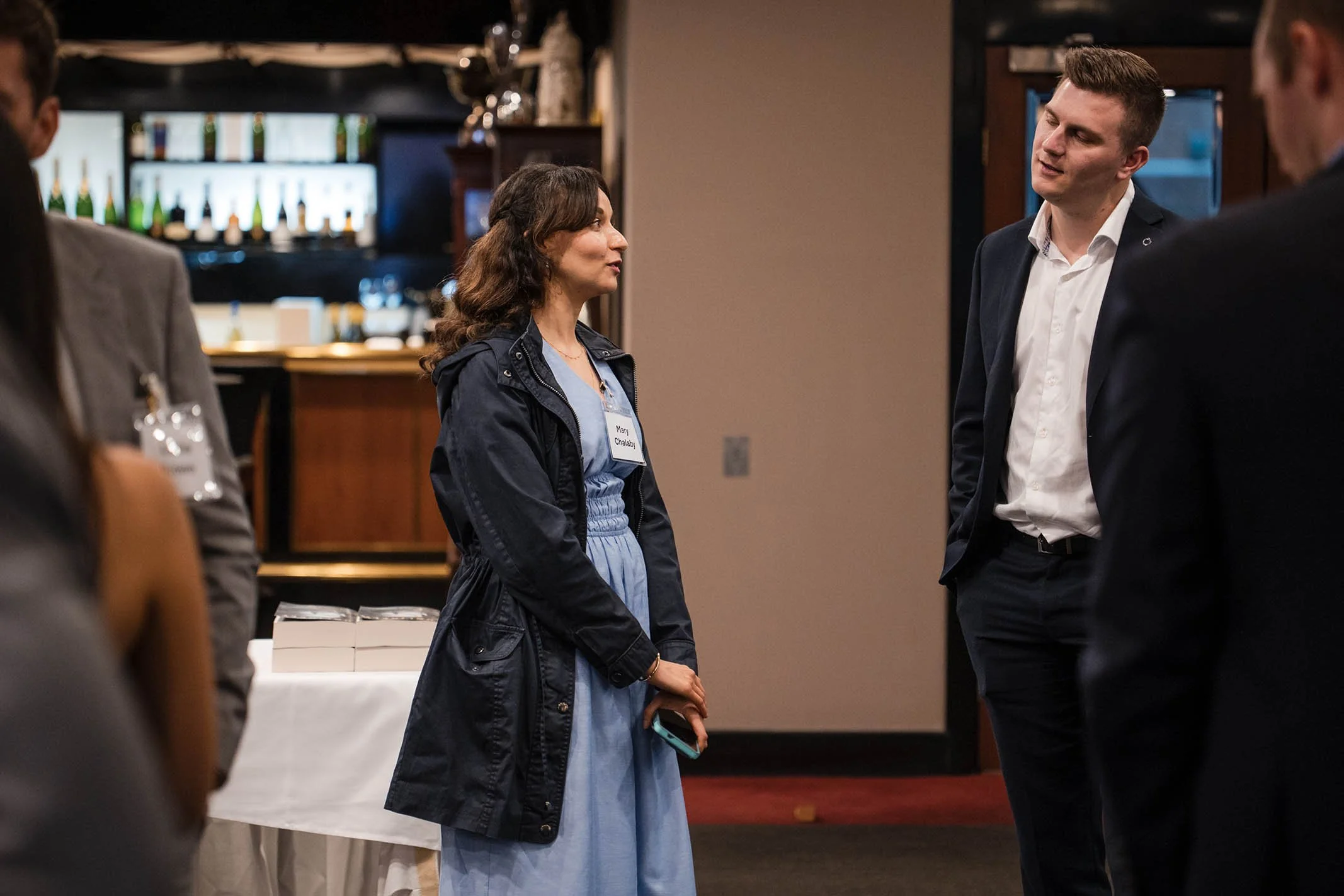 A woman talking to two men at a social event in a restaurant or lounge setting with a bar in the background.