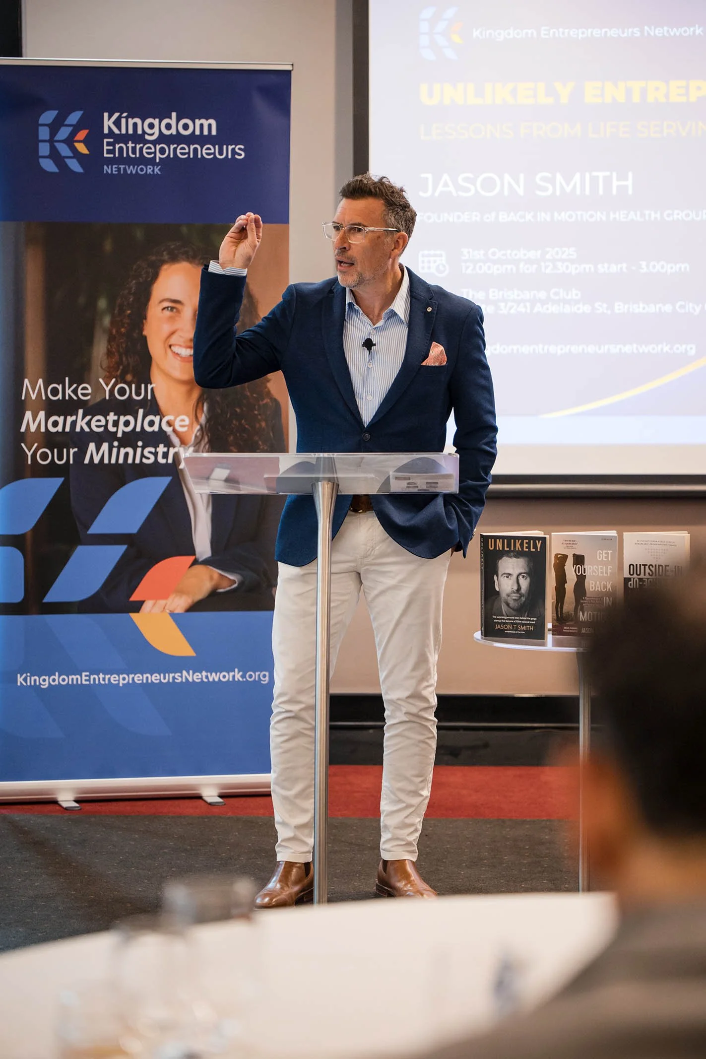 A man in a blue blazer, striped shirt, and beige pants giving a speech at a Kingdom Entrepreneurs Network event, standing at a clear podium with books displayed on a table beside him. A large screen behind him shows the event details and his name, Ja