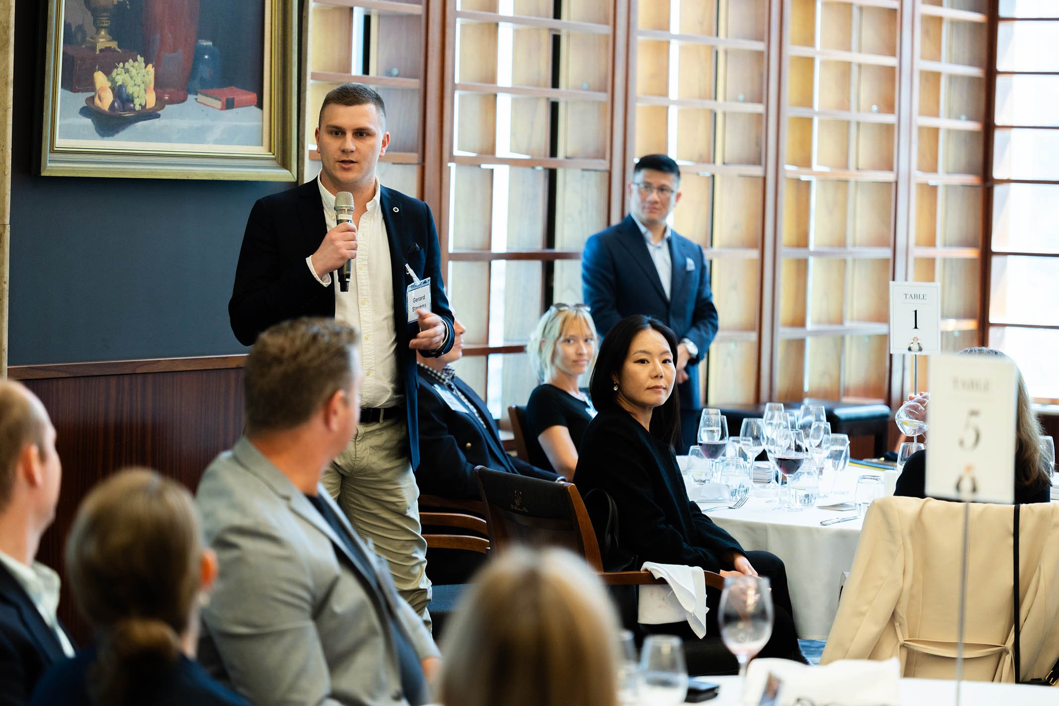 A man in a suit holding a microphone speaks at a formal event with seated attendees at tables with wine glasses, some looking at him and others at the camera. There are two men standing behind him, a woman sitting at the table in the foreground. The 