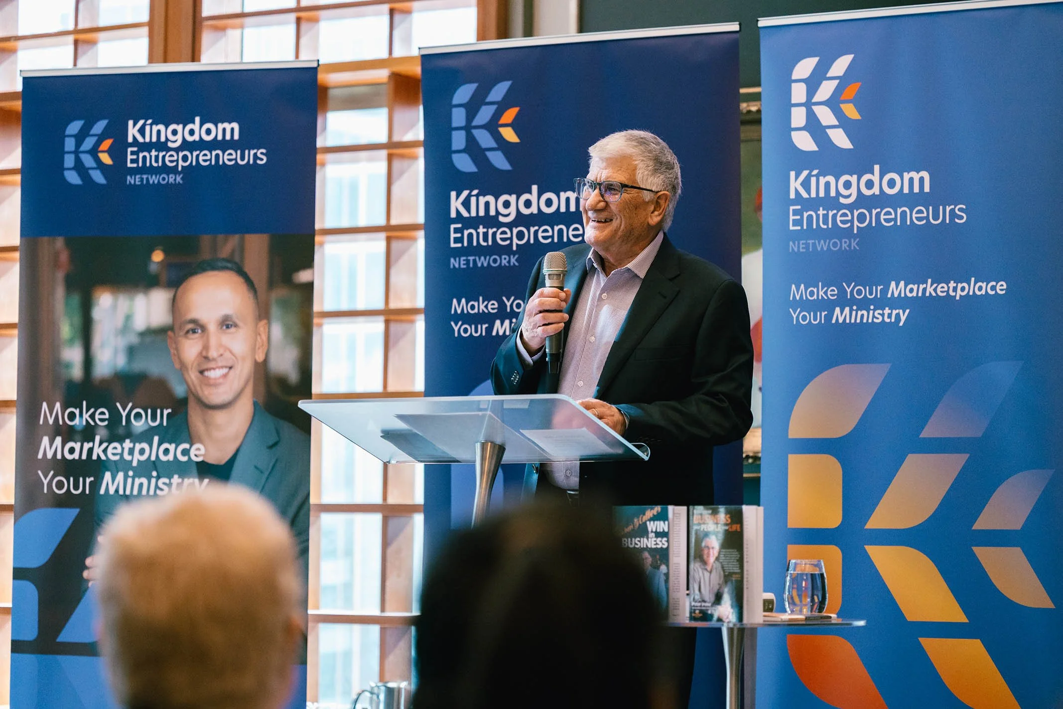 A man in a suit and glasses speaking at a podium with a microphone at a Kingdom Entrepreneurs Network event. Behind him are blue banners with the organization logo and the message 'Make Your Marketplace Your Ministry.'