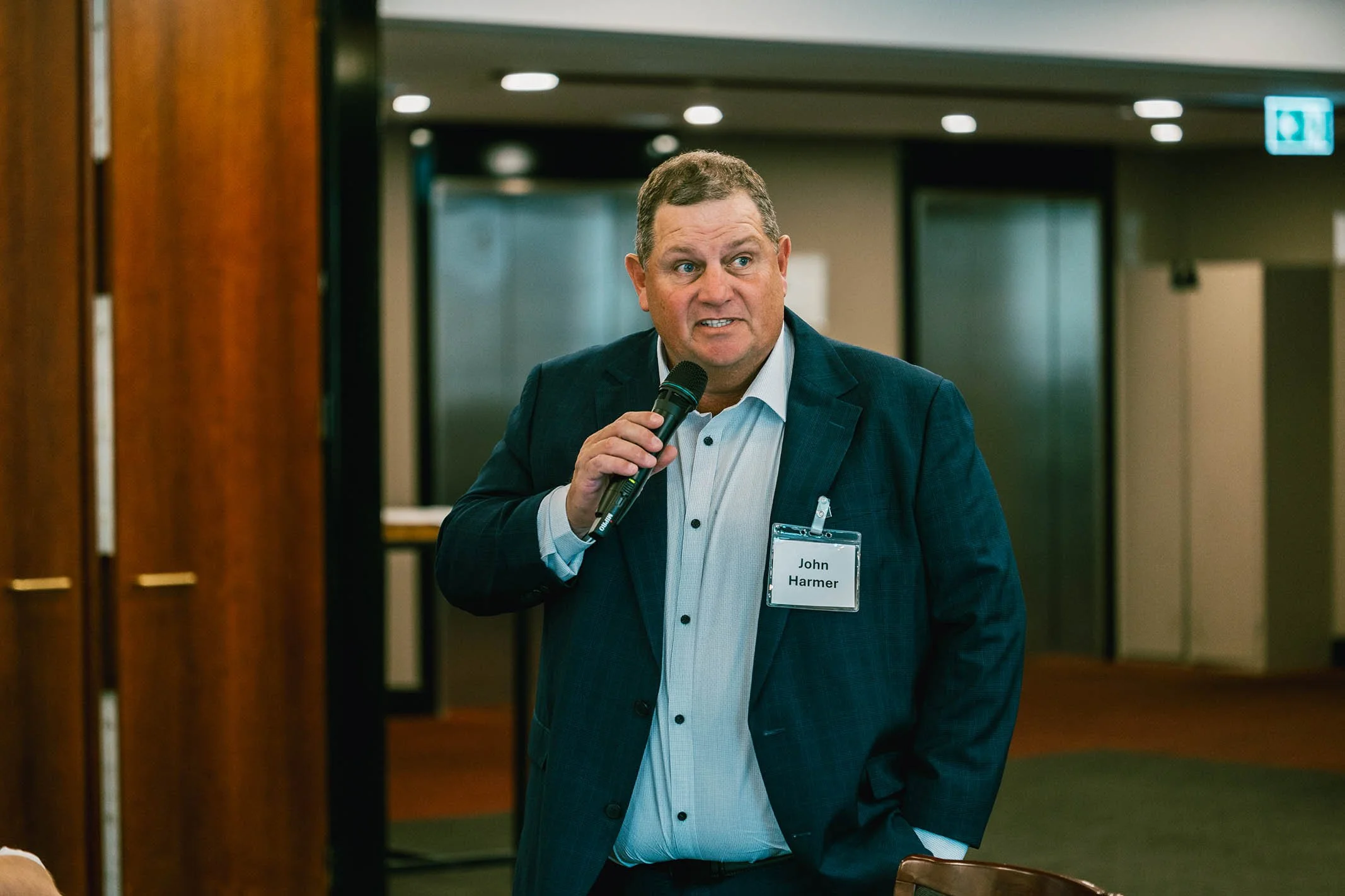 A man in a suit holding a microphone, standing indoors at an event, wearing a name tag that reads 'John Harmer'.