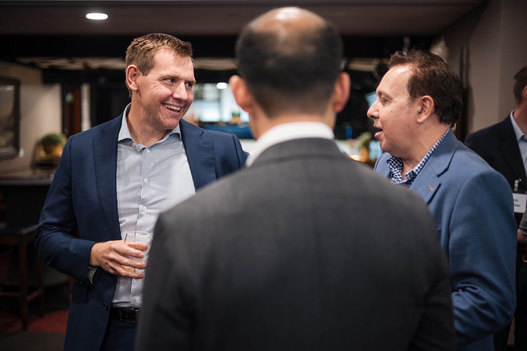 Three men in business attire engaged in a conversation at a social event, with one man holding a drink and smiling.