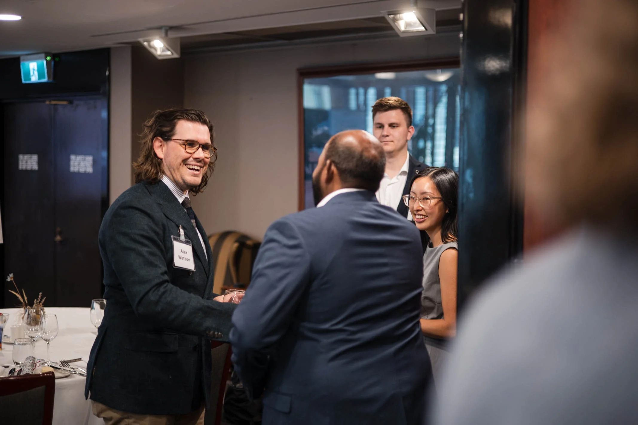 Group of people engaged in conversation at a formal event, with two men and two women smiling and talking near a table with glassware.