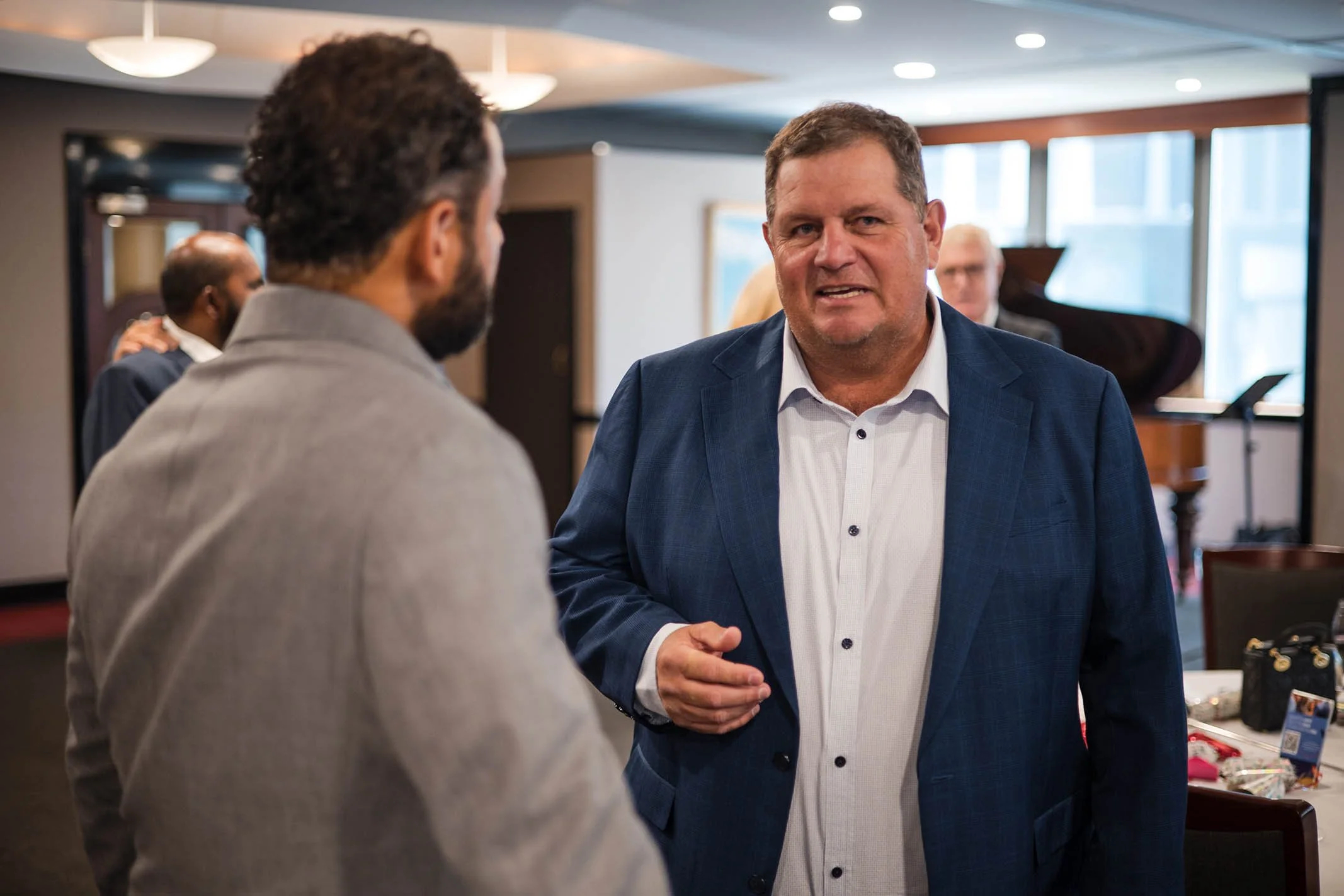 Two men engaged in a conversation at a social event, with a third person in the background and a grand piano visible in the venue.