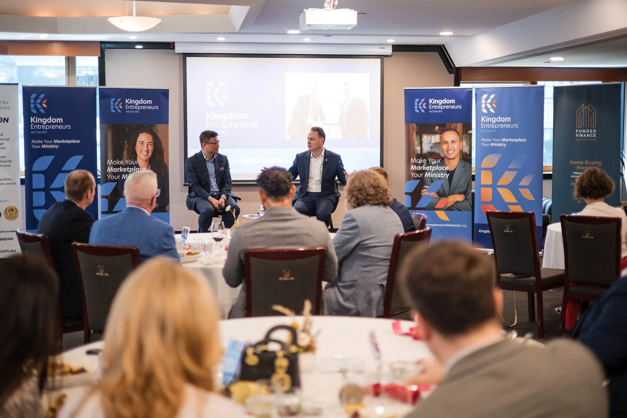 A business conference with three speakers on stage, two men and one woman, and an audience of diverse professionals seated at round tables. Banners with 'Kingdom Entrepreneurs' and related messages are displayed around the stage.