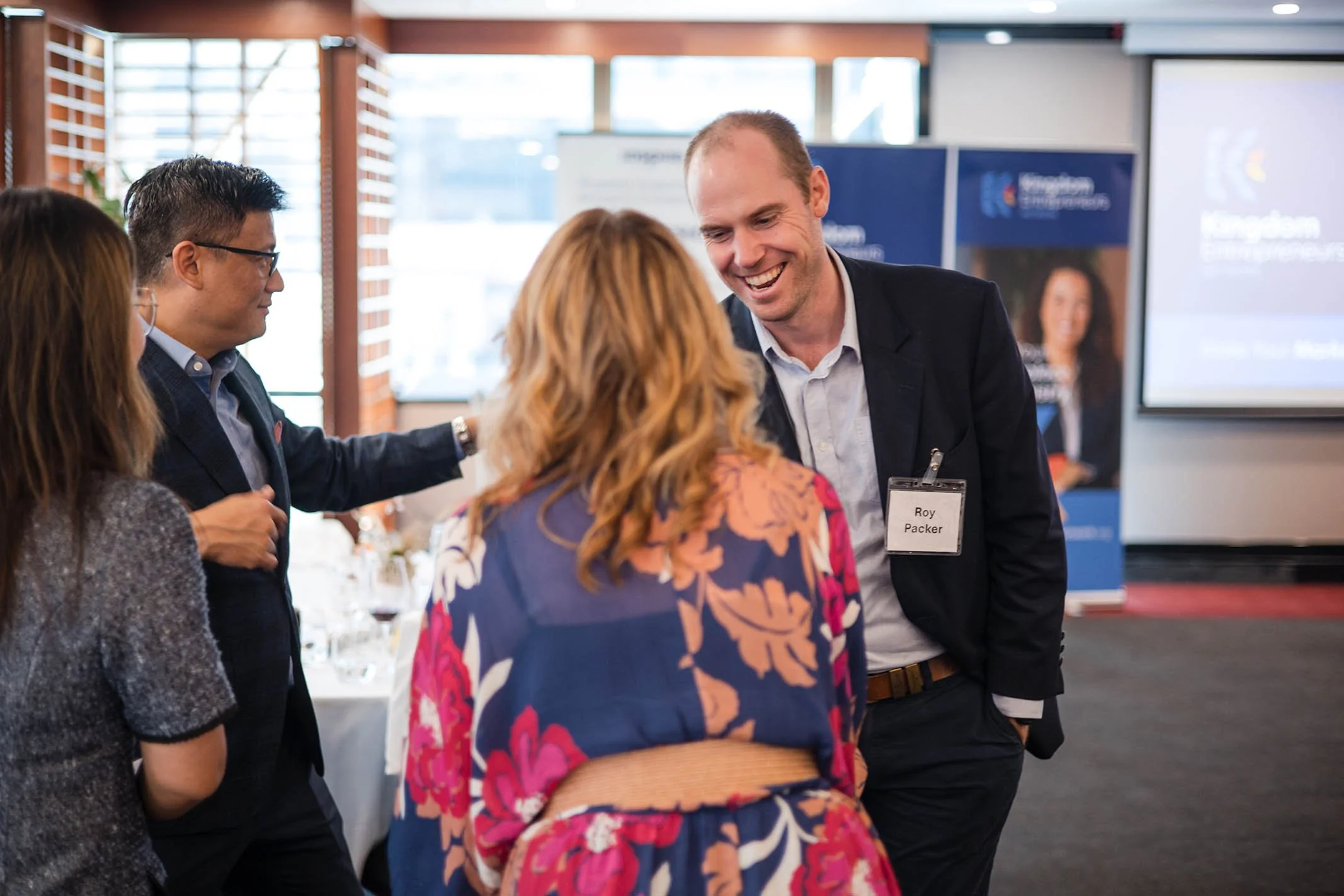 A group of people talking at a professional event with a man smiling and wearing a name badge, in a room with large windows and presentation screens.