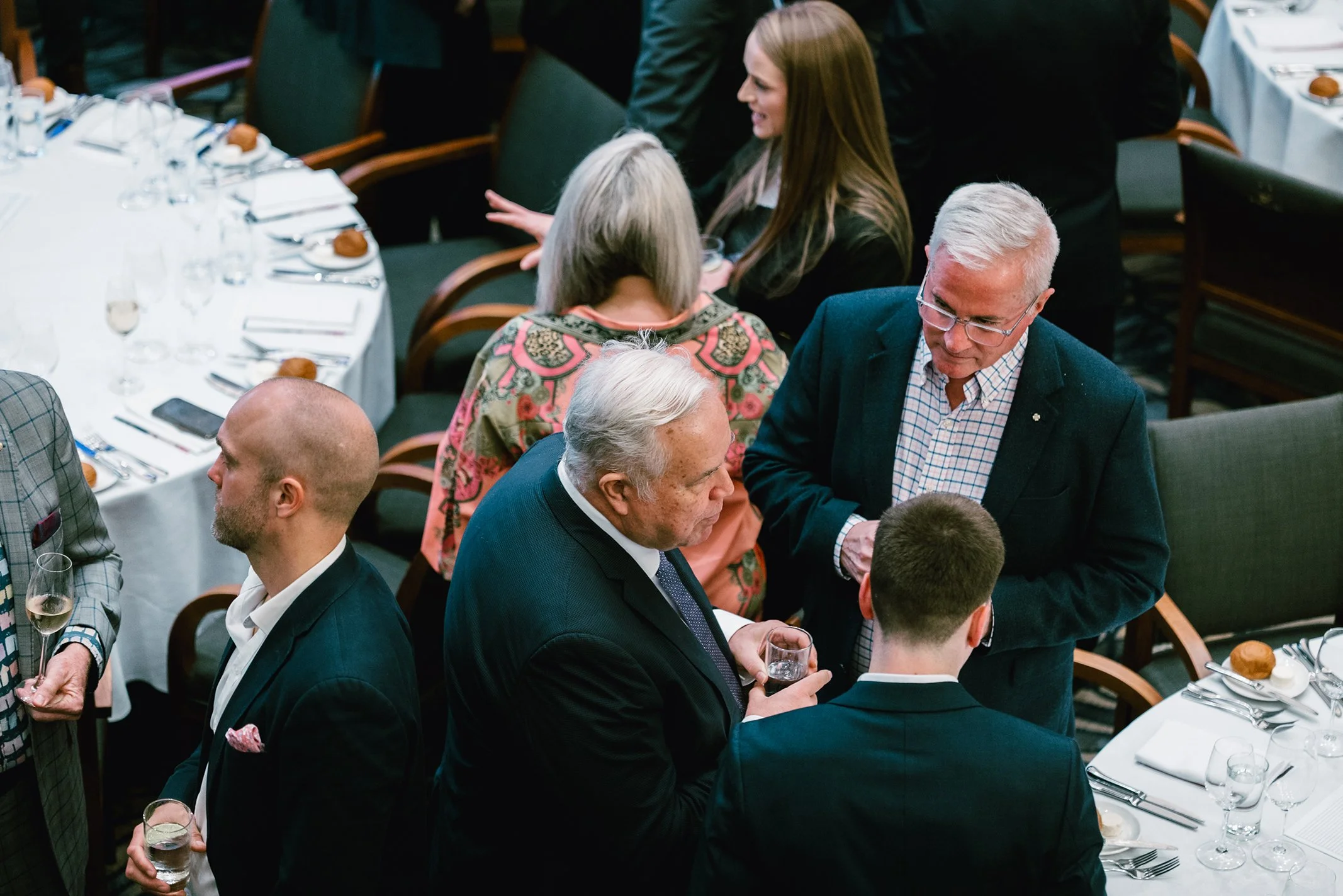 People socializing at a formal dinner, standing and chatting around tables set with wine glasses, silverware, and bread rolls.
