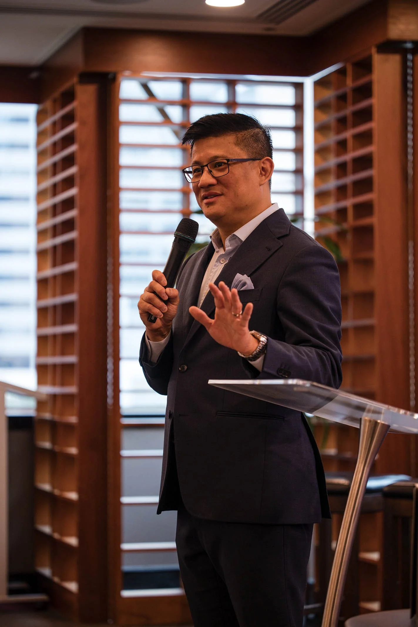 A man in a dark suit and glasses holding a microphone and speaking at a wooden podium in a room with wooden shelves and large windows.
