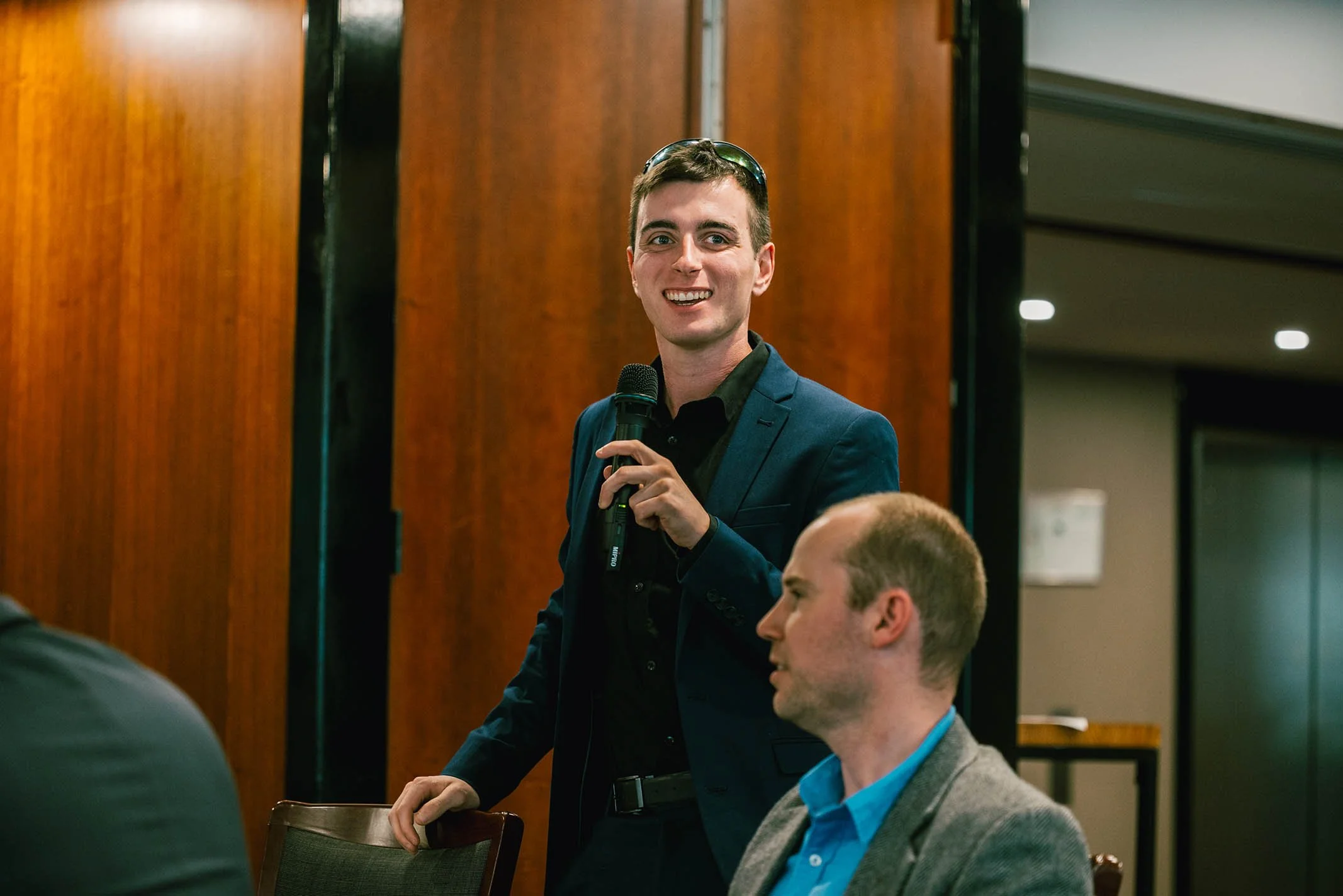 A young man in a dark suit and black shirt holding a microphone, standing and speaking at a conference or meeting, with other seated men around him.