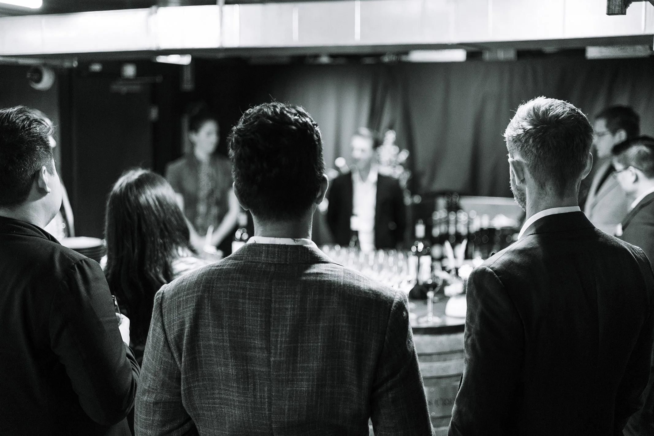 A group of people dressed in formal attire observing a speaker at an event in a dimly lit venue.