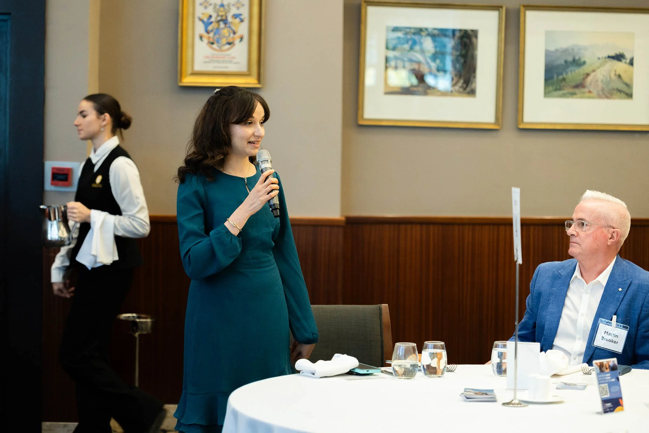 A woman in a teal dress holding a microphone speaking to a seated man in a blue blazer at a restaurant or banquet setting.