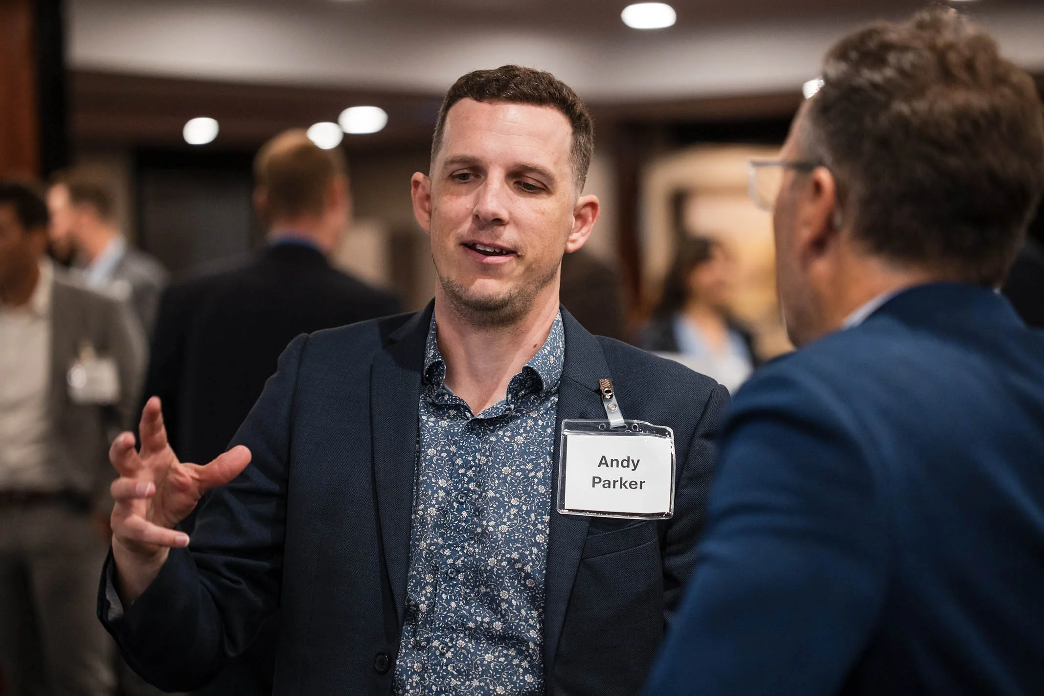 A man named Andy Parker with short dark hair, wearing a navy blazer and floral shirt, is talking to another man at a professional event, with blurred people and conference room background.