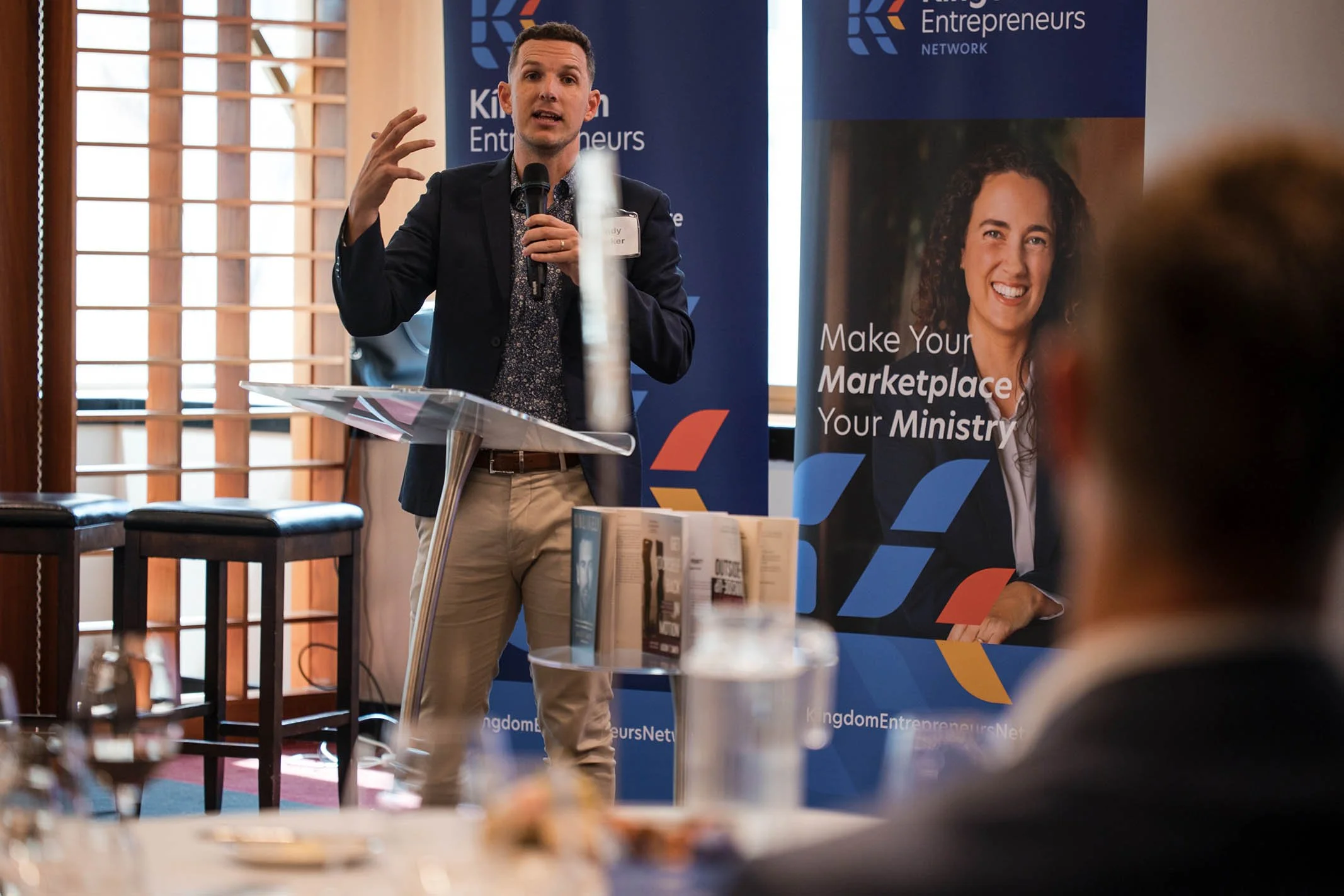 A man in a dark blazer and light-colored pants is speaking into a microphone at a podium, gesturing with one hand. Behind him, there are two banners with the logo and text for 'Kingdom Entrepreneurs Network' and a smiling woman. The scene appears to 