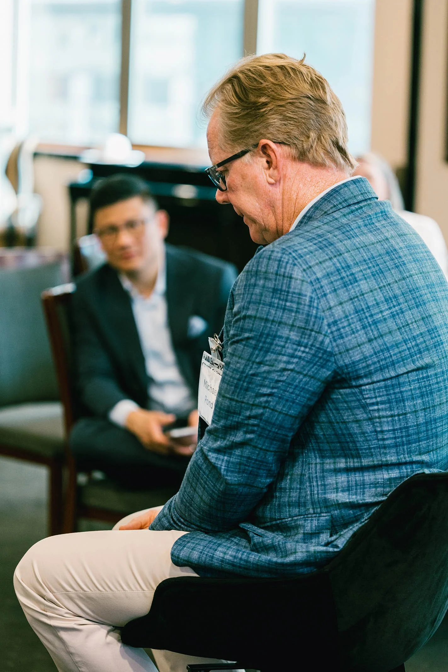 Two men in business attire at a professional event or conference, one sitting and looking down, the other with a name tag, sitting on a chair in an indoor setting.