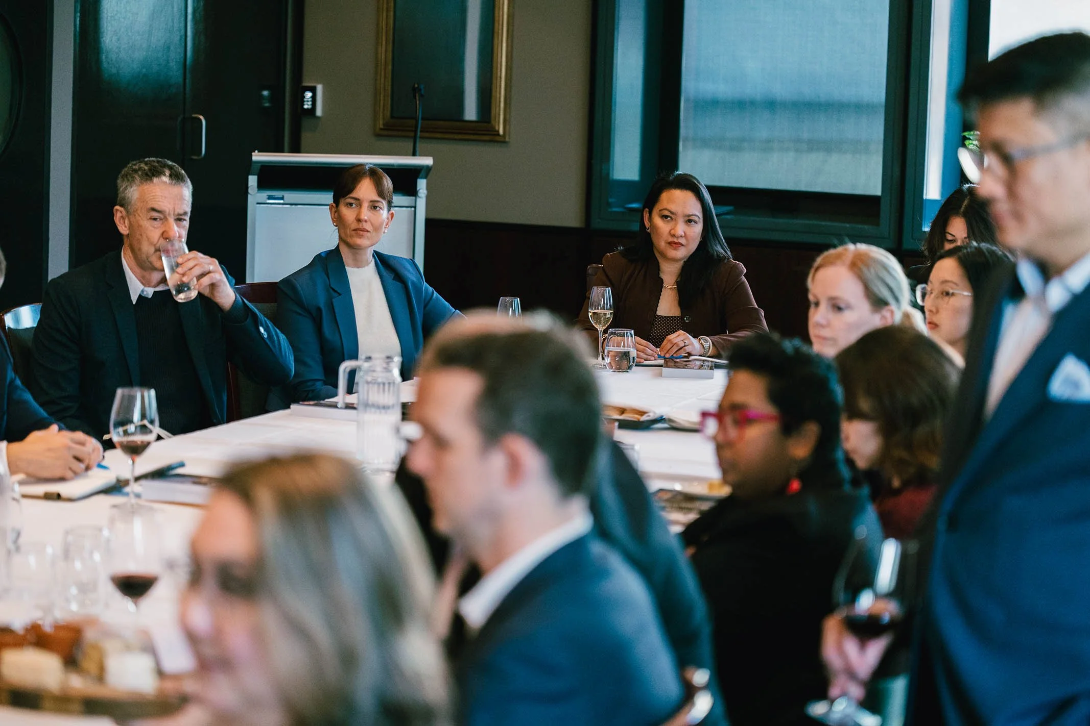 A group of diverse people attending a business meeting or conference, sitting around a table with glasses of water and wine, in a formal setting.