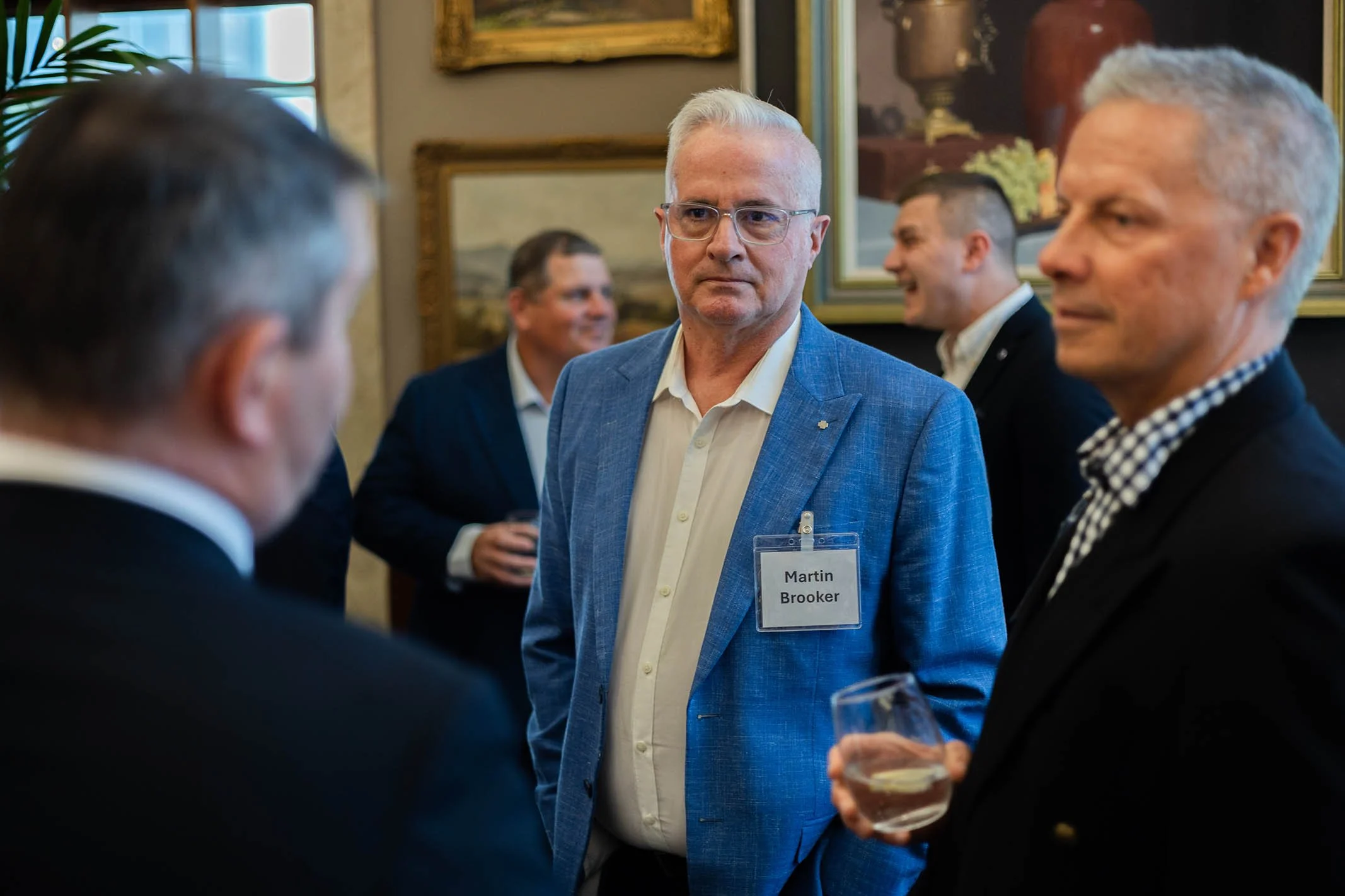 A group of men at a professional networking event, engaged in conversation. One man, with glasses and a name tag reading 'Martin Brooker,' is in focus, wearing a light blue blazer. Others are seen in the background, holding drinks, with paintings on 
