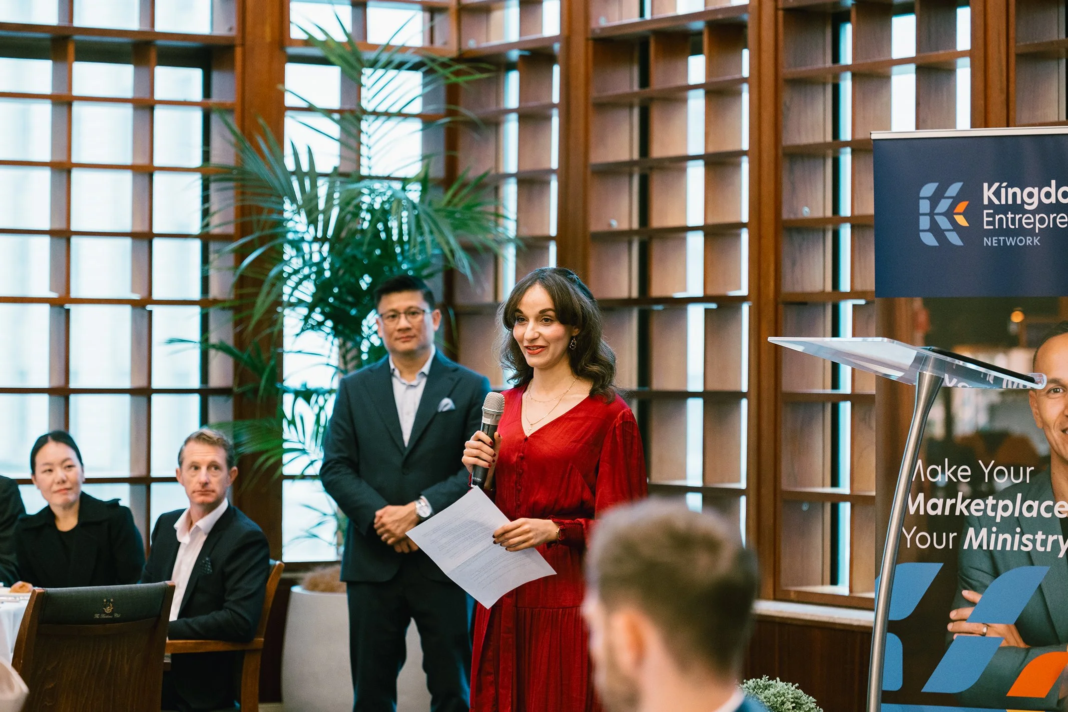 A woman in a red dress holding a microphone and a sheet of paper, speaking at a formal event in a room with wooden paneling and large windows. There are seated and standing people listening, and a blue sign reading 'Kingdom Entrepreneur Network' with