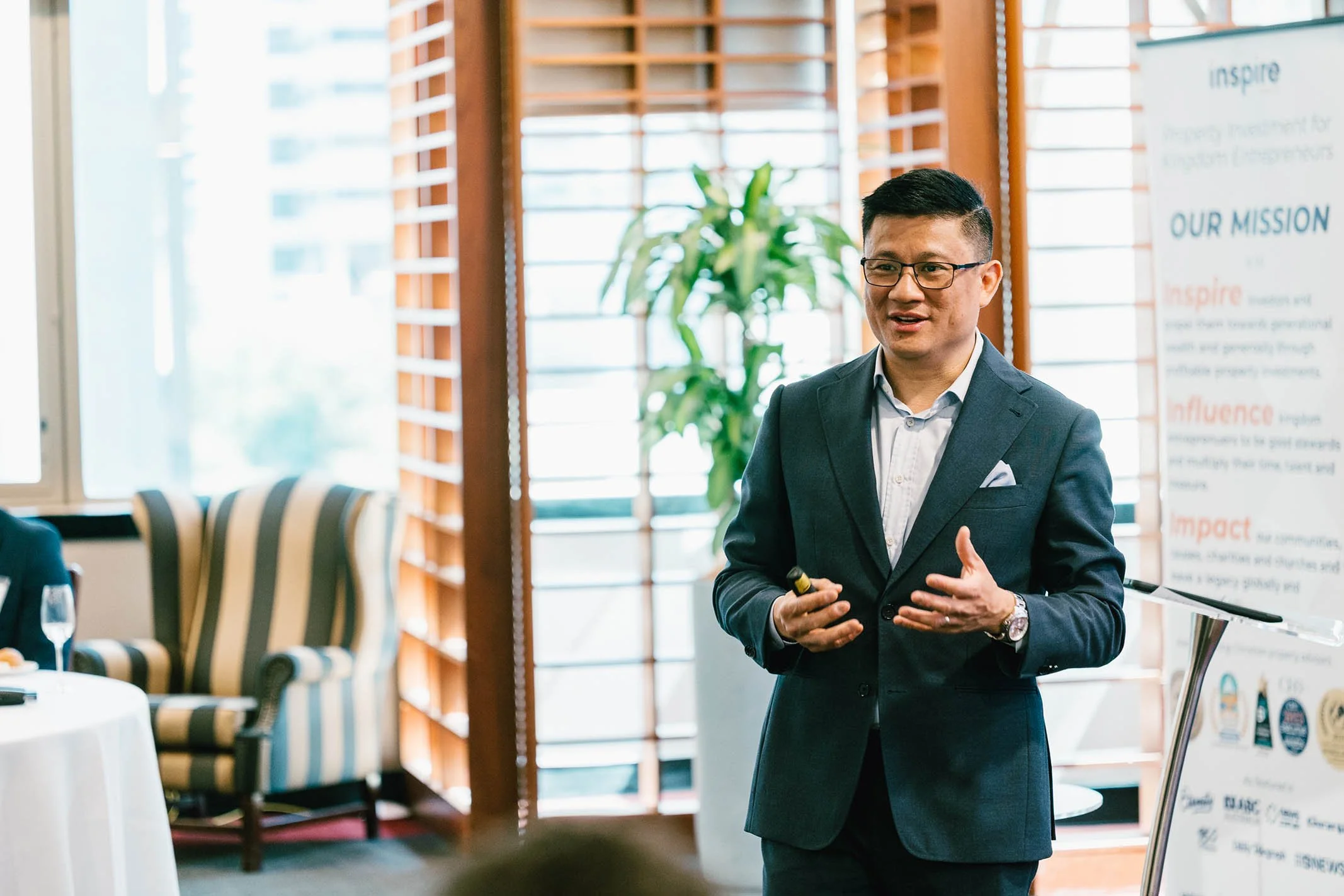A man in a dark suit and glasses giving a presentation in a bright, wooden-paneled room with large windows and a poster in the background.