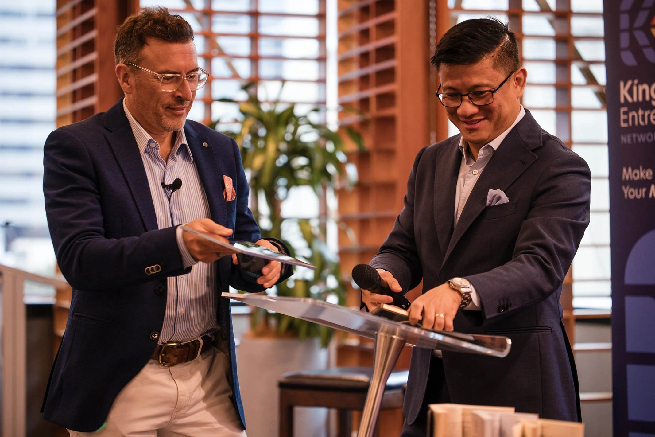 Two men in business suits standing at a podium, with one holding a microphone and the other looking at a tablet, in a conference setting.