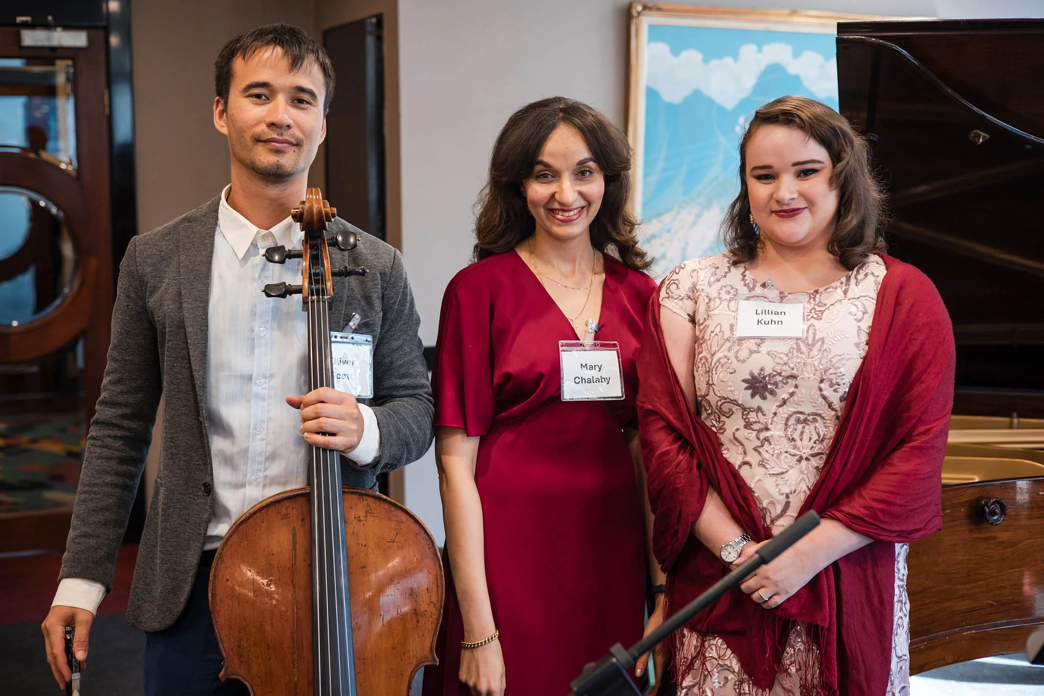 Three people standing together at a formal event, with one person holding a cello, all wearing name tags, and a grand piano visible in the background.
