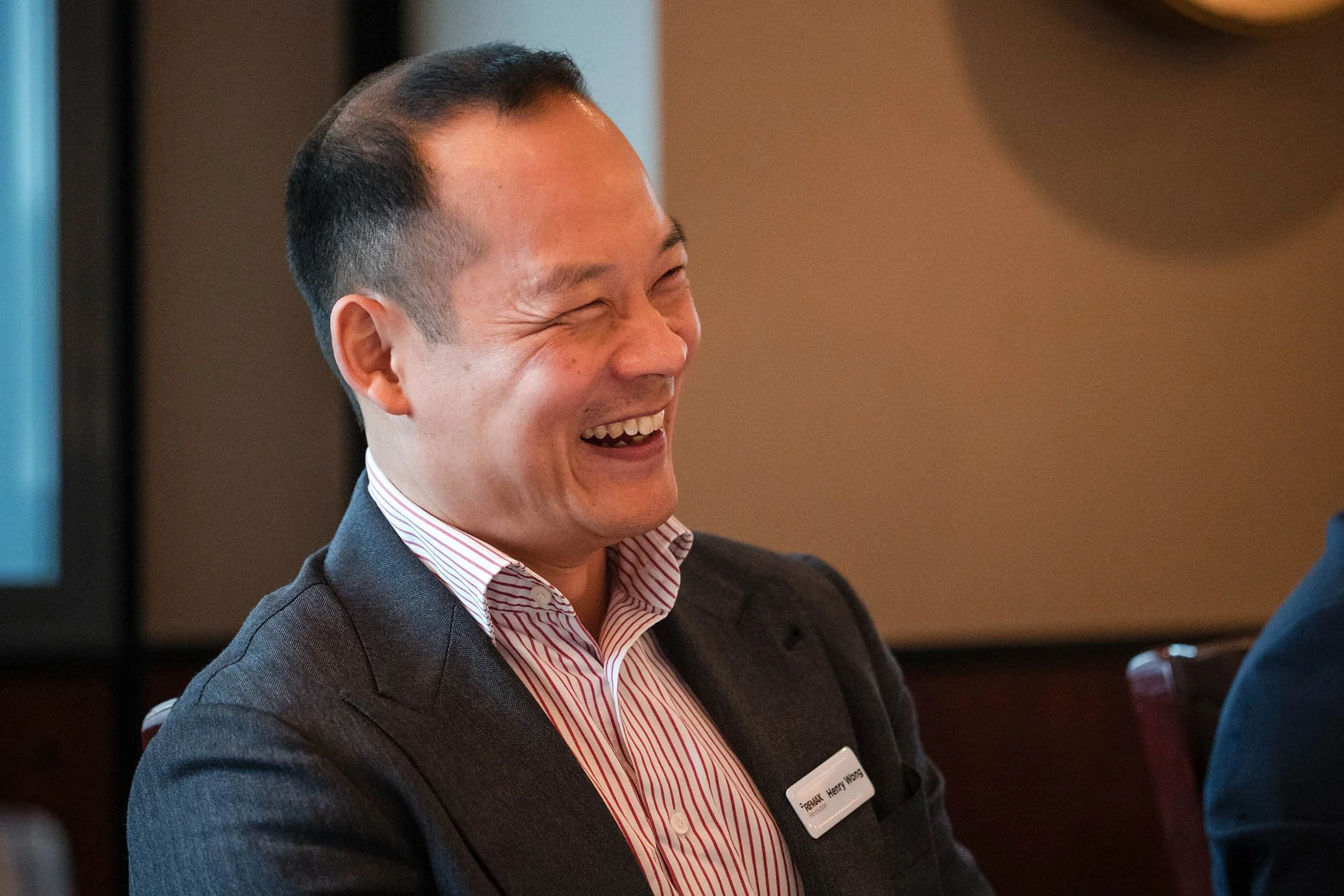 A man with short black hair, wearing a dark blazer and a name tag, is laughing and smiling, sitting at a table in an indoor setting.