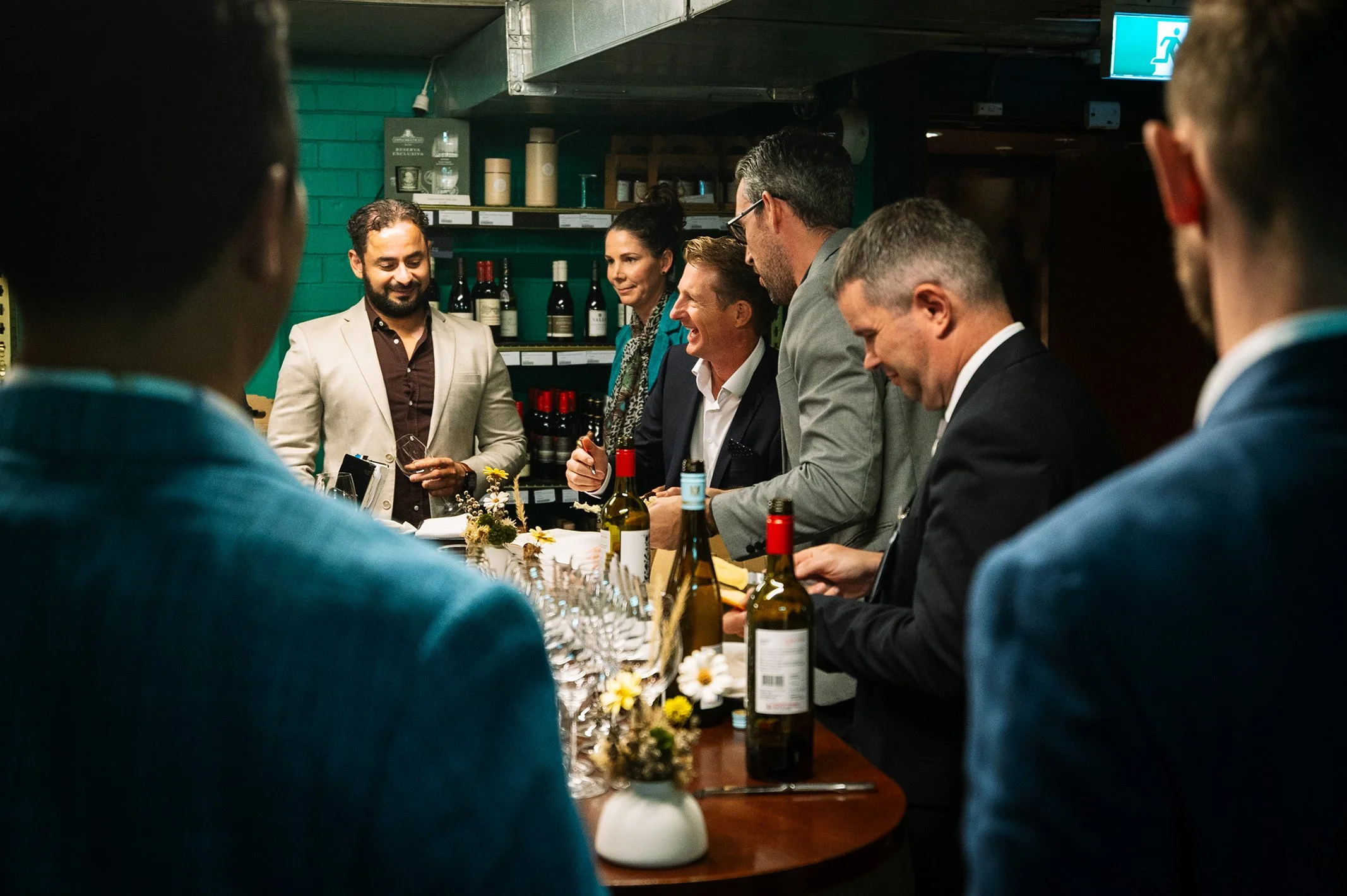 A group of people at a wine tasting event, standing around a table with wine bottles and glasses, engaging in conversation with a presenter.