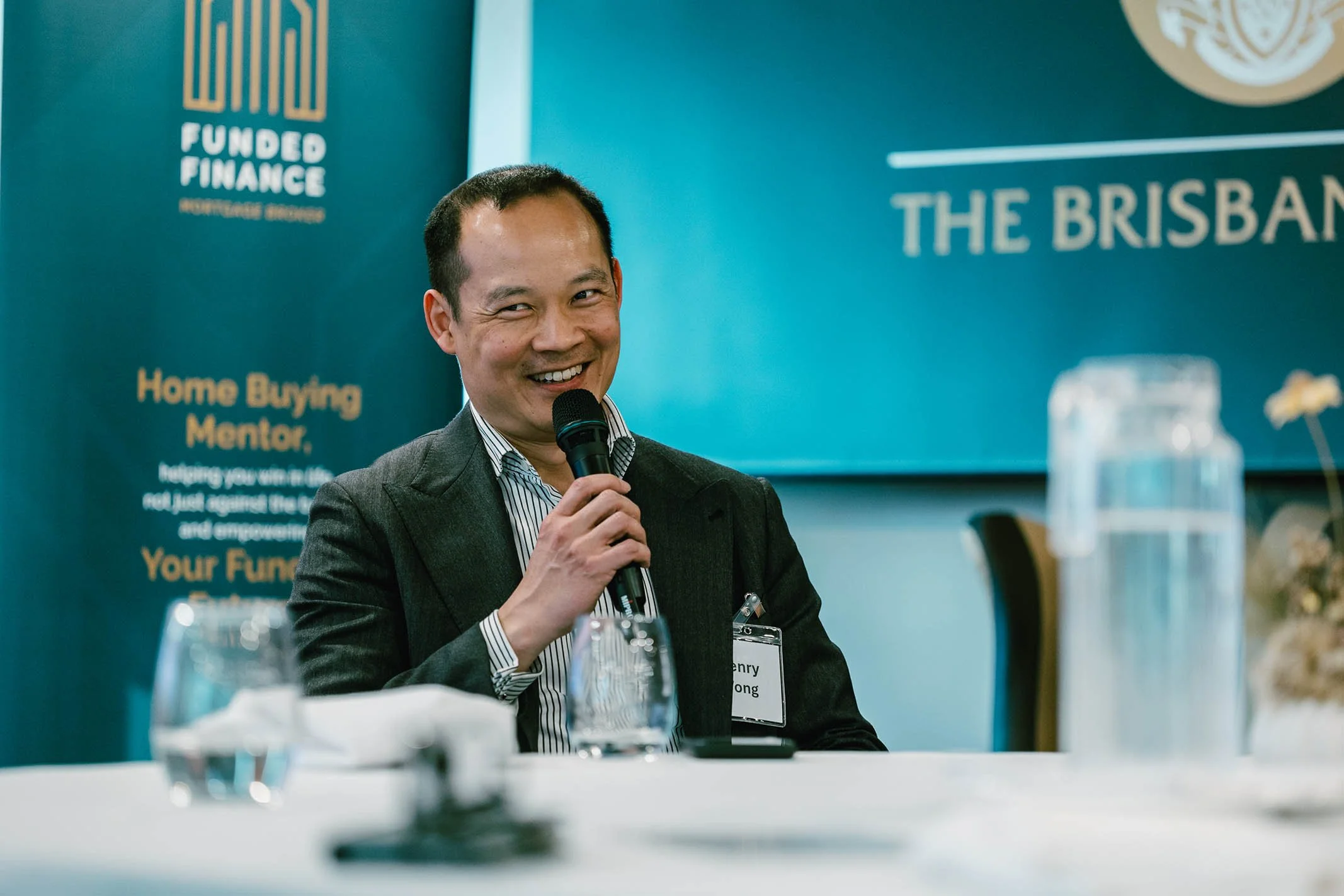 A man in a suit speaking into a microphone at a conference, with a blue backdrop displaying the words 'Funded Finance' and 'The Brisbane'.
