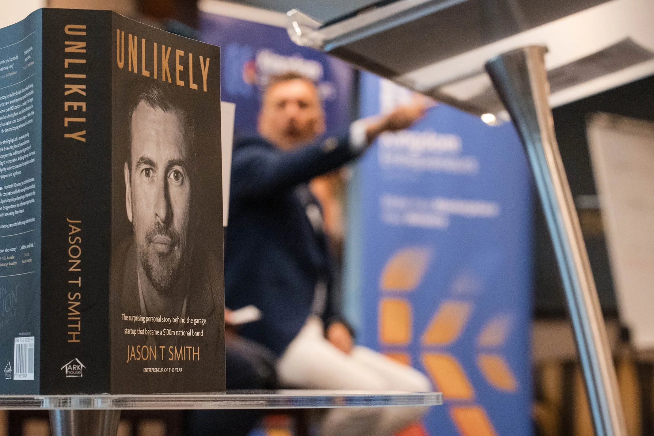 A book titled 'Unlikely' about Jason T Smith, with a black and white portrait of him on the cover. The background shows a man in a blue jacket and white pants sitting at a table.