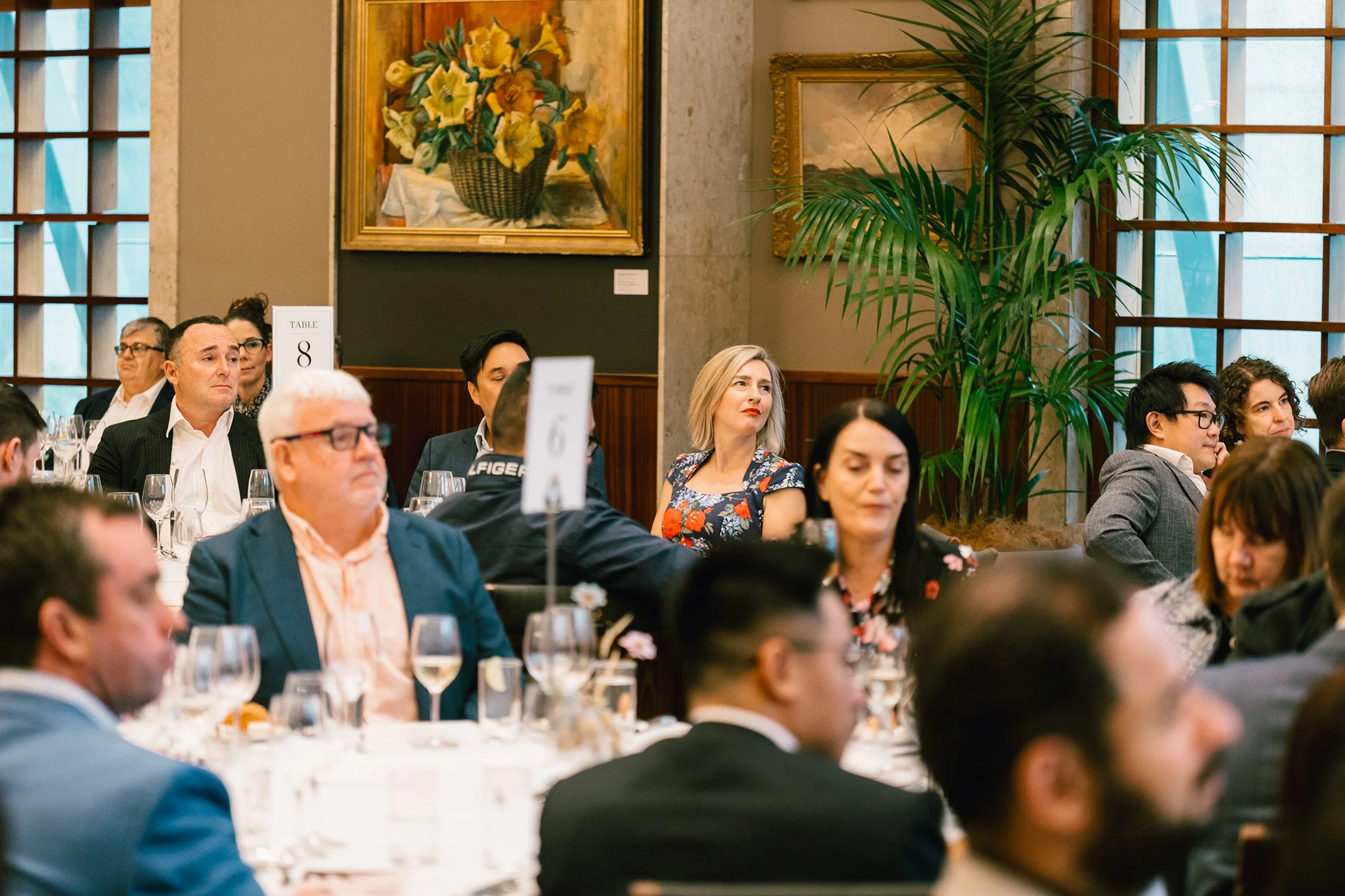 People attending a formal event seated at round tables with white tablecloths and glassware, in a room with large windows, framed paintings, a green potted plant, and modest decor.