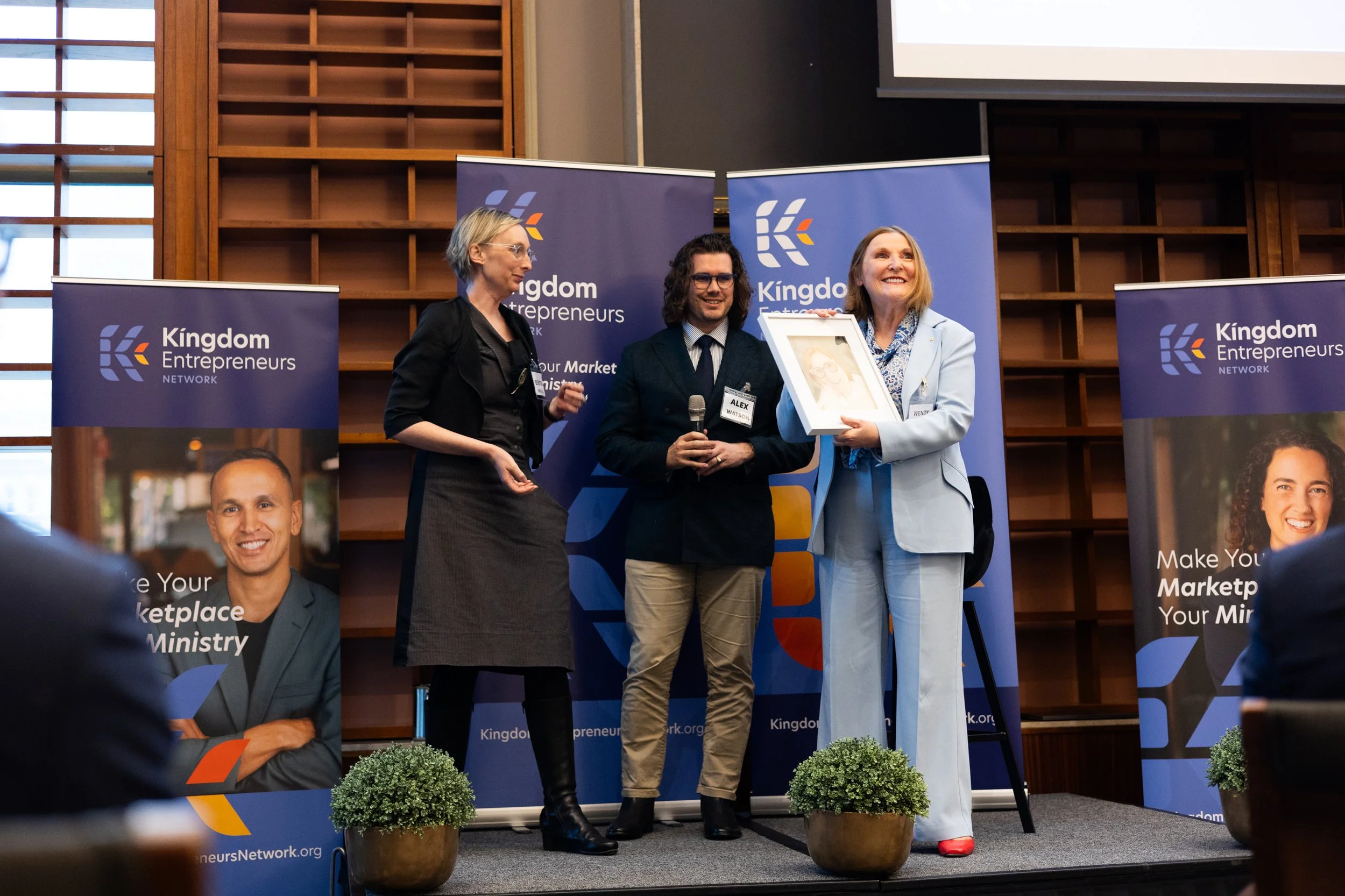 Three individuals are on a stage at a Kingdom Entrepreneurs Network event, with two women and a man. The woman on the right is presenting a framed photo to the woman on the left, who is smiling. The man in the middle is holding a microphone. The back