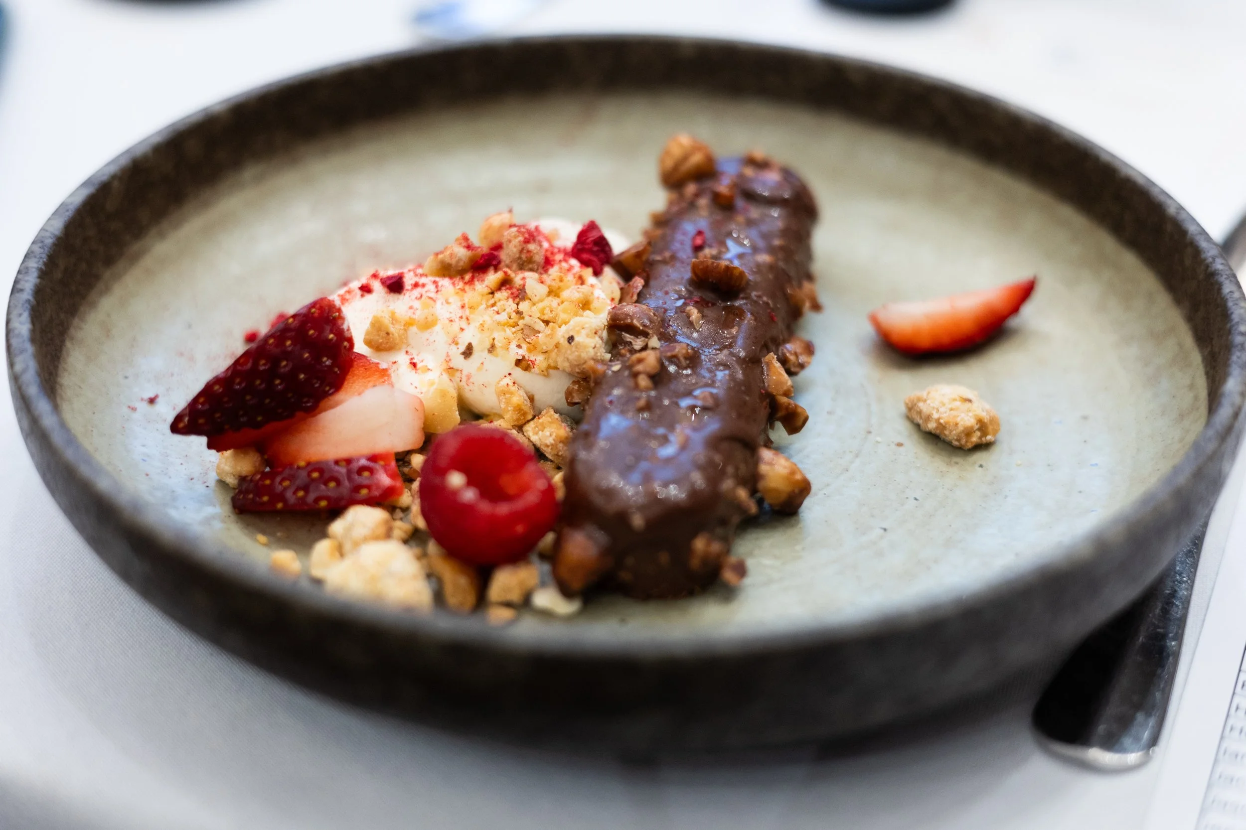 Dessert of chocolate-coated banana, fresh strawberries, raspberries, whipped cream, granola, and nuts served on a gray ceramic plate.