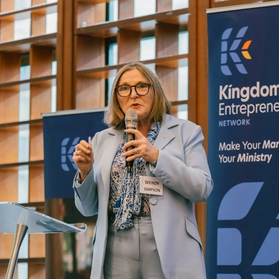 A woman with glasses and shoulder-length hair speaking into a microphone at a conference, standing next to a banner that reads 'Kingdom Entrepreneurship Network'.