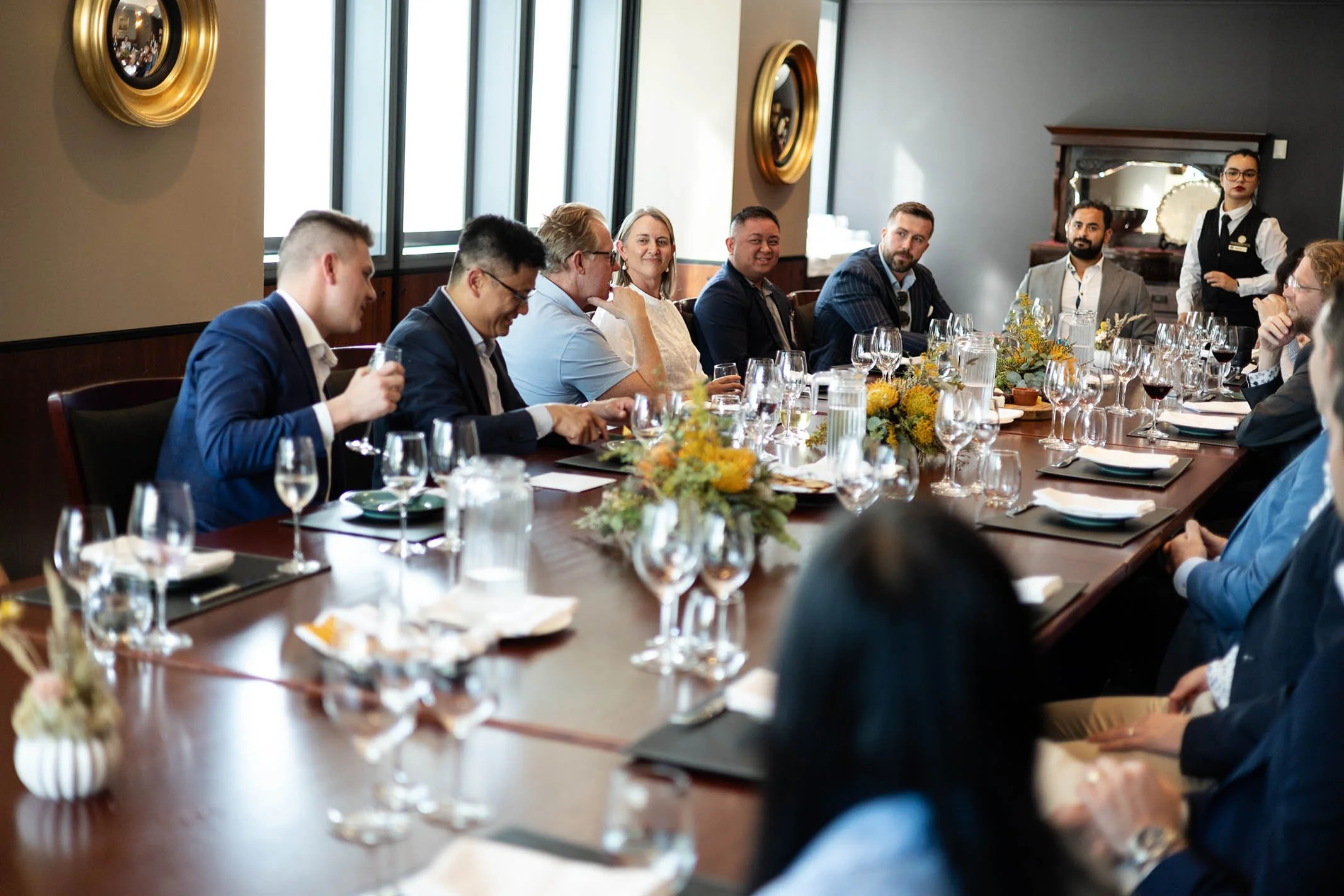 A group of people in formal attire seated around a large dining table during a meeting or celebration, with floral centerpieces and glassware on the table.