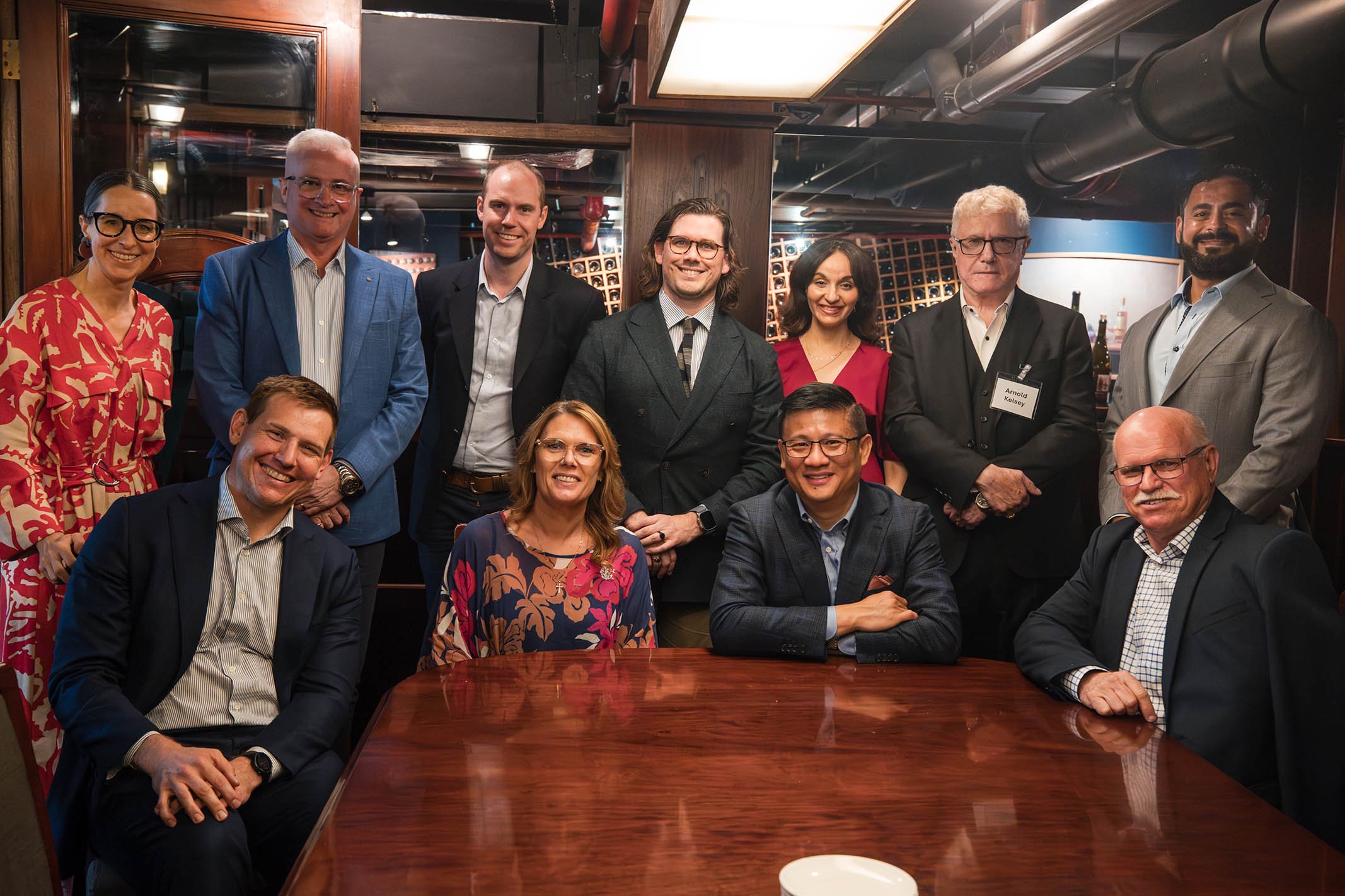 A group of eleven professionally dressed people gathered around a wooden table in a restaurant or private dining room, with some standing in the background and some sitting in the foreground.