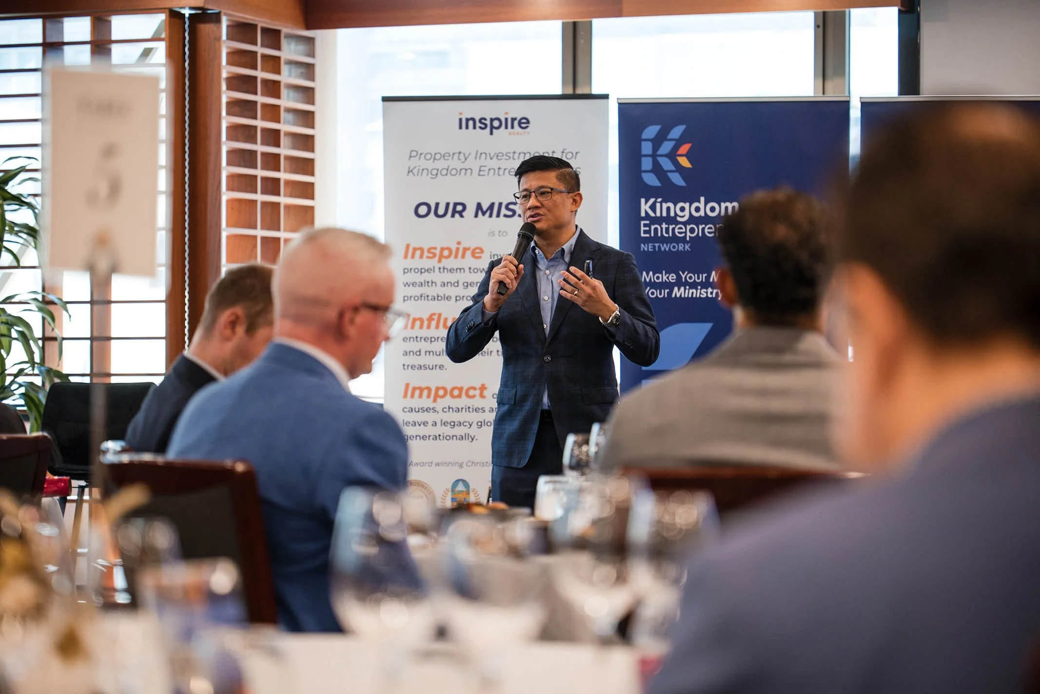 A man in a suit speaking into a microphone during a conference, with a backdrop of banners related to Kingdom Entrepreneurship Network, and several attendees seated at tables in the foreground.