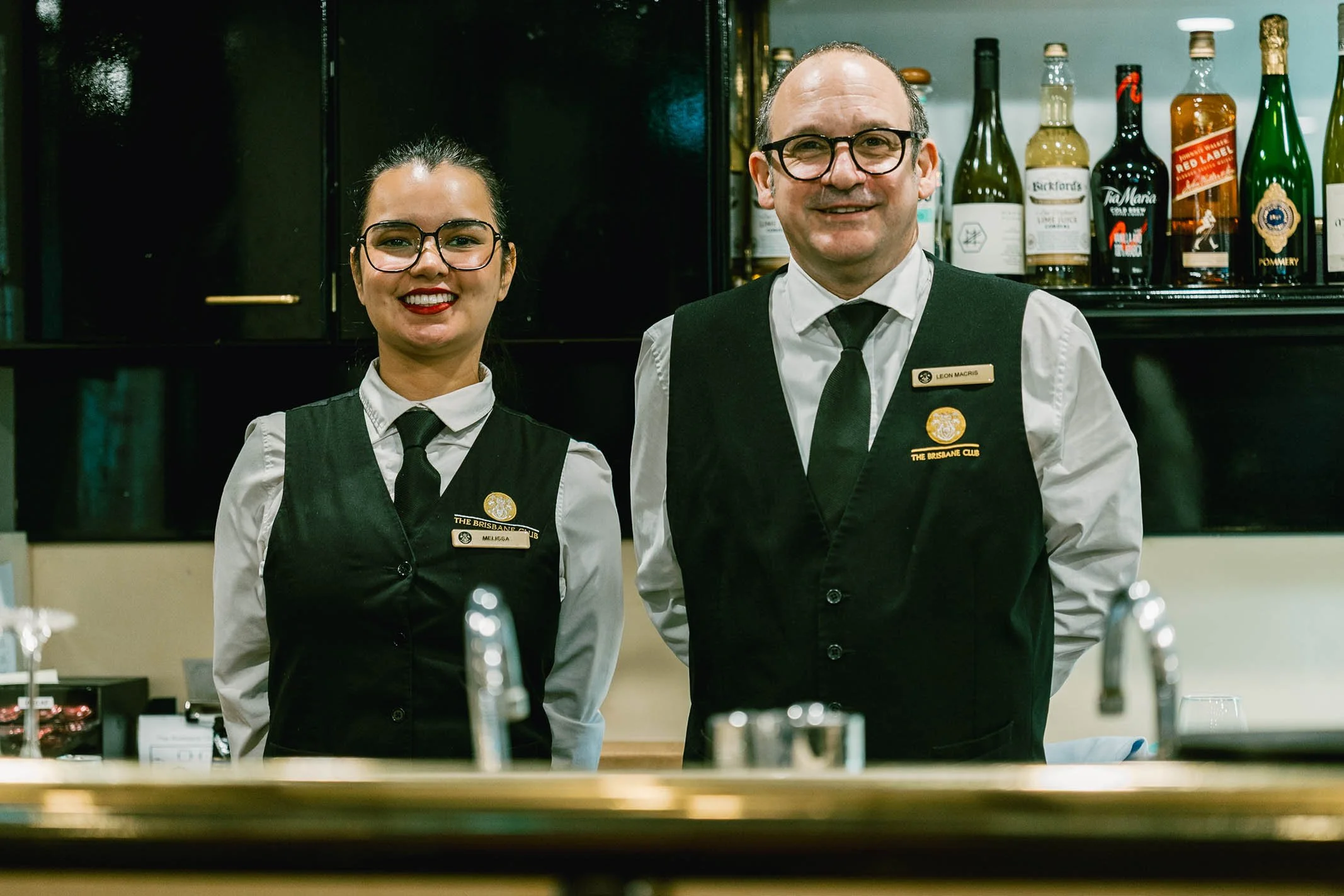 Two bartenders, a woman and a man, standing behind a bar in a pub, smiling and wearing professional black vests and white shirts with ties, with bottles of alcohol on shelves behind them.