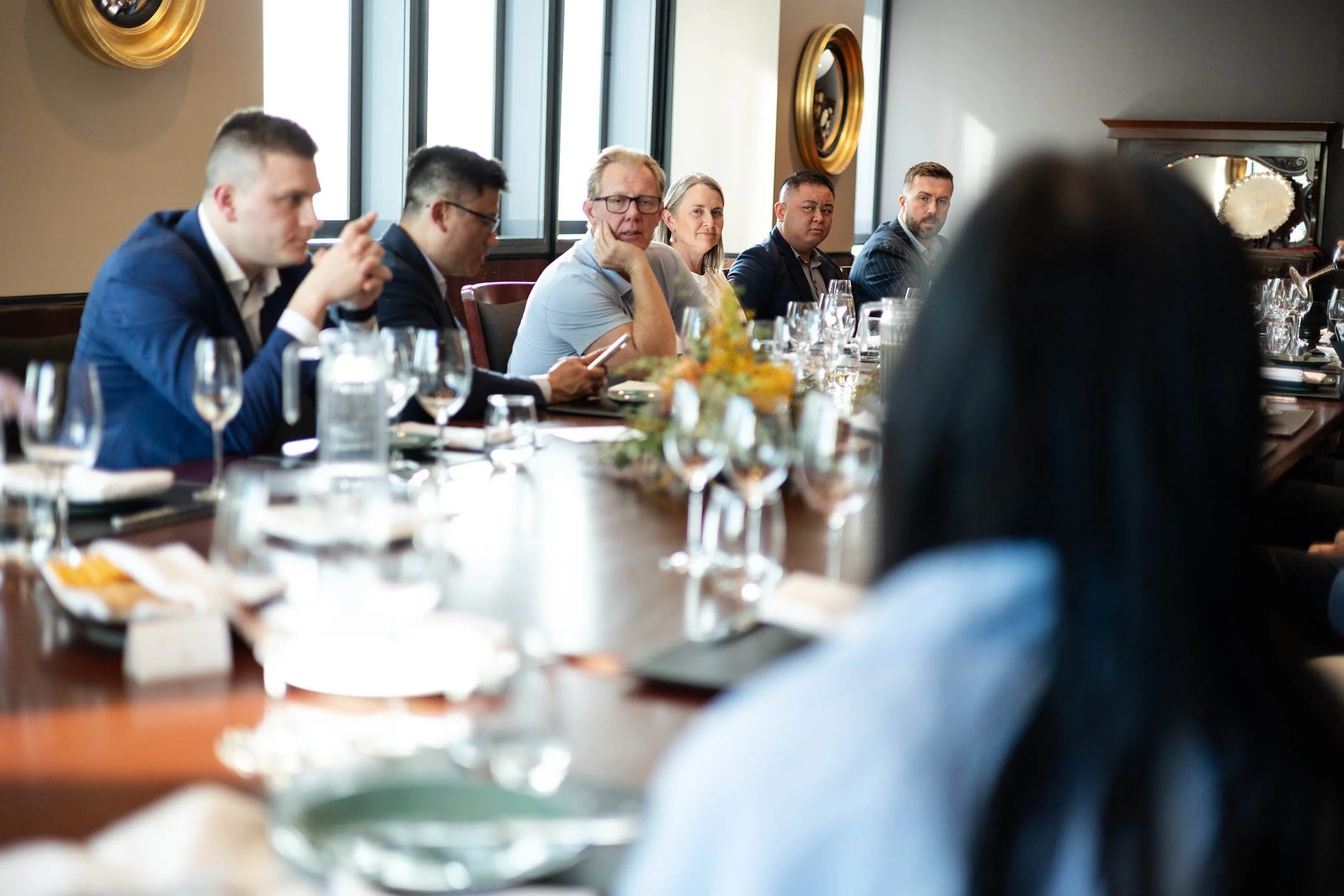 A group of people seated around a long conference table in a meeting room, some looking at their phones or at the speaker, with glasses and plates on the table.