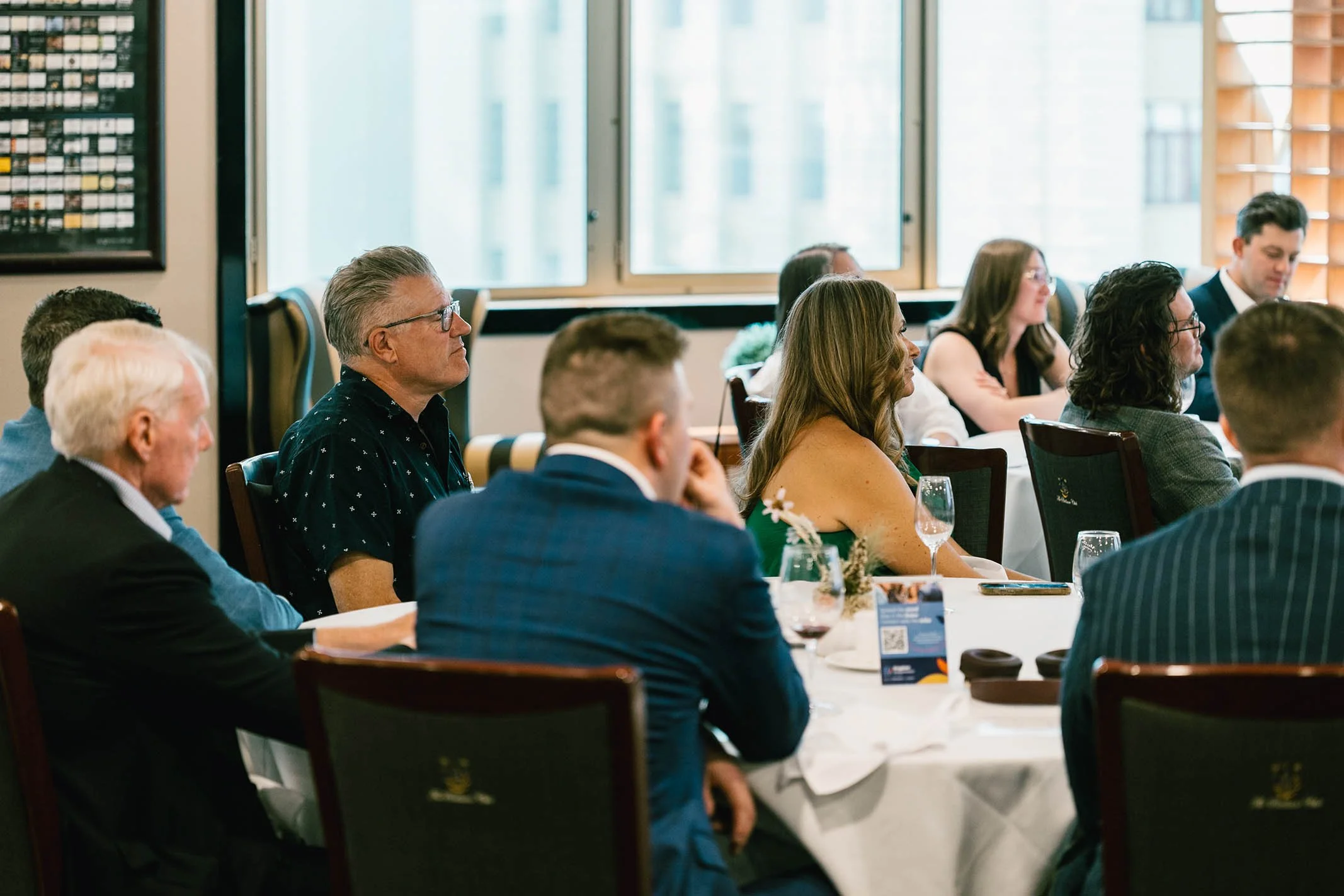 A group of people attending a conference or meeting in a bright room with large windows, sitting at tables with glasses of wine and documents.